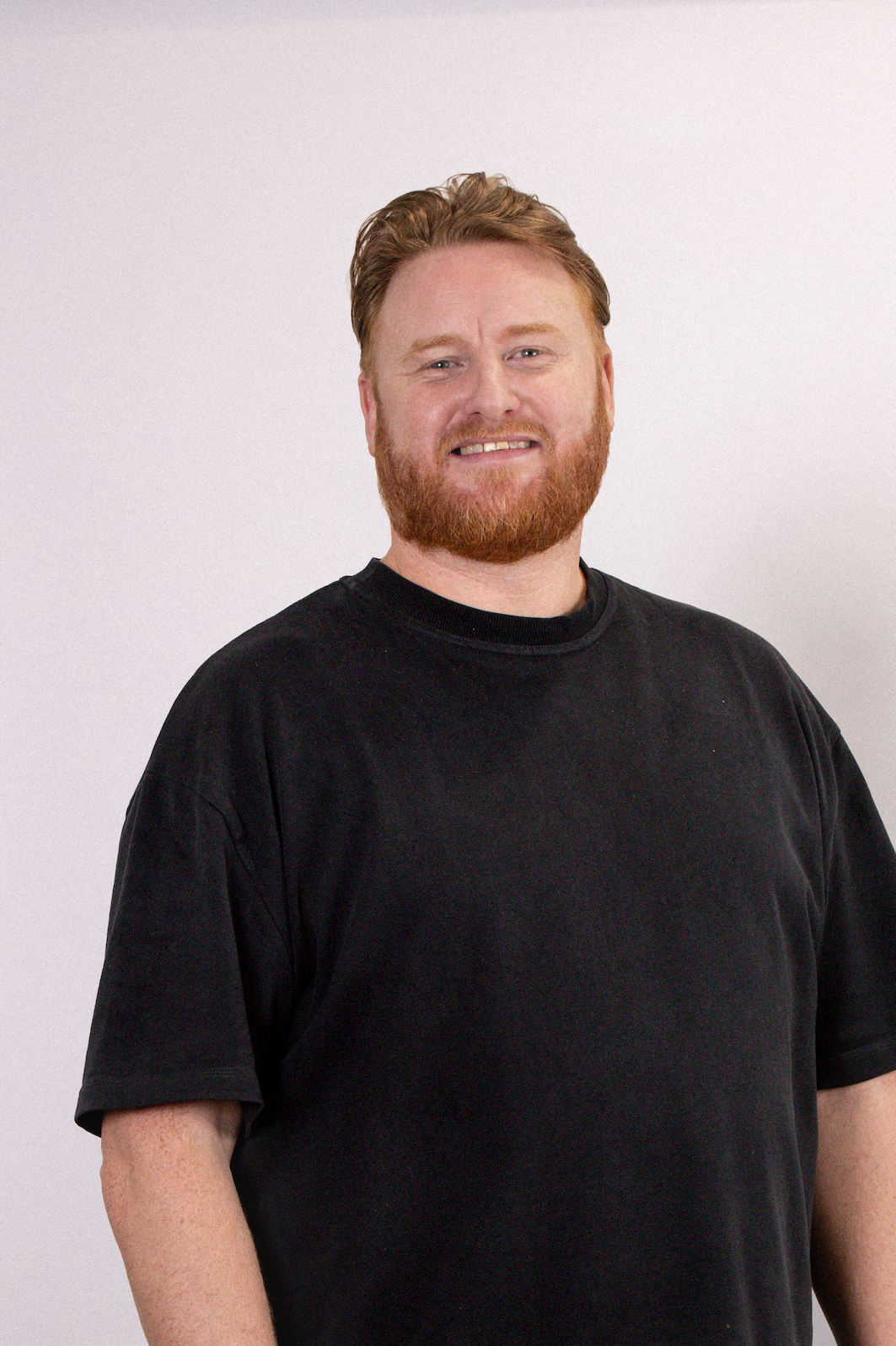 A man with red hair and a beard smiling, wearing a black T-shirt, standing against a plain white background.