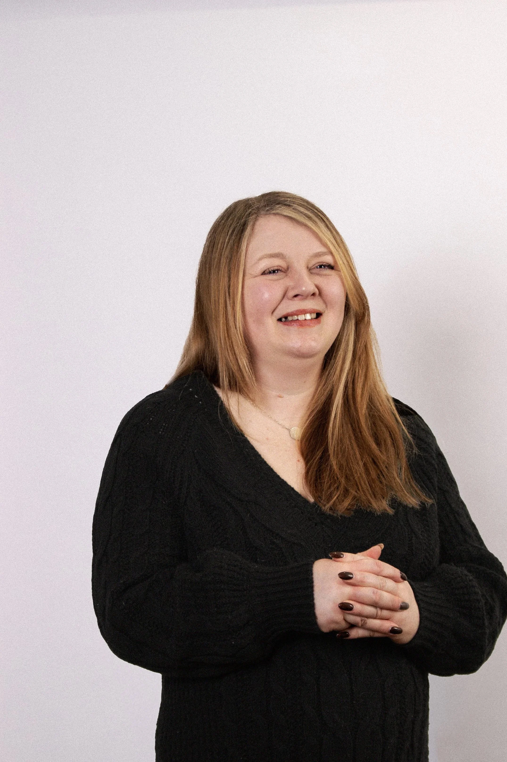A woman with long red hair, smiling, wearing a black sweater, standing against a plain white background.