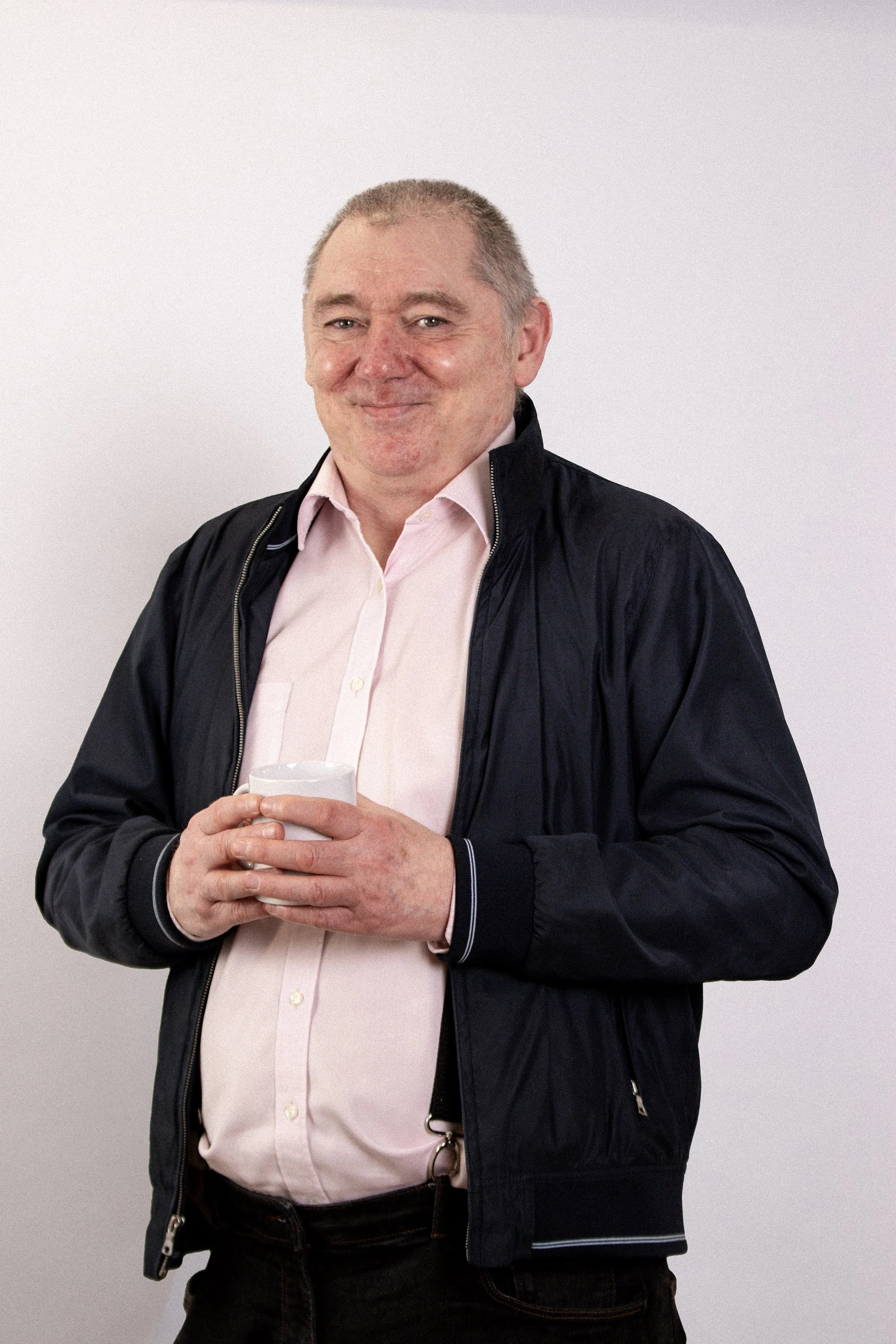 A  man with short gray hair, wearing a light pink shirt and a black jacket, holding a glass of water, standing against a plain white background and smiling.