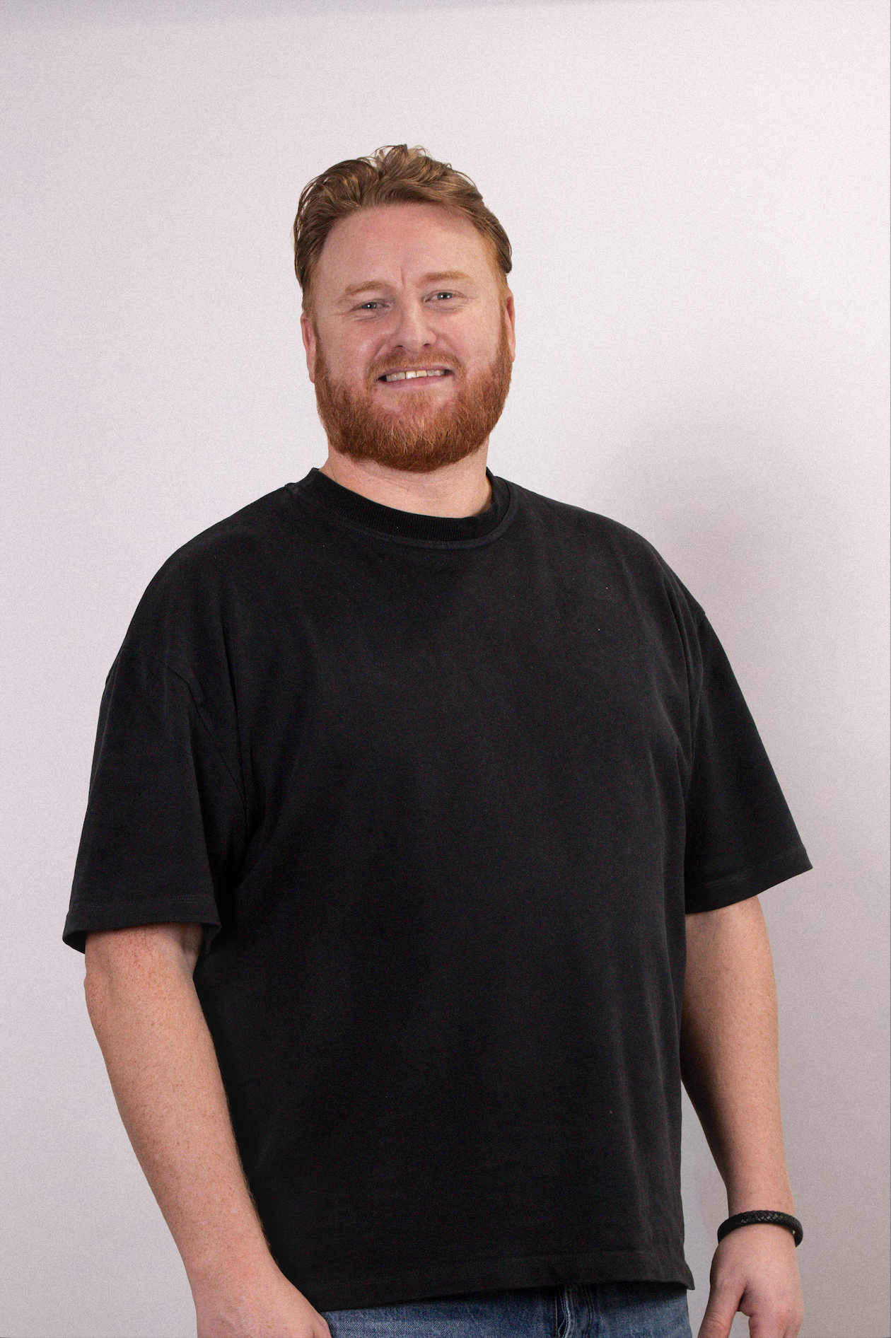 A man with red hair and beard wearing a black t-shirt and jeans standing against a white background.