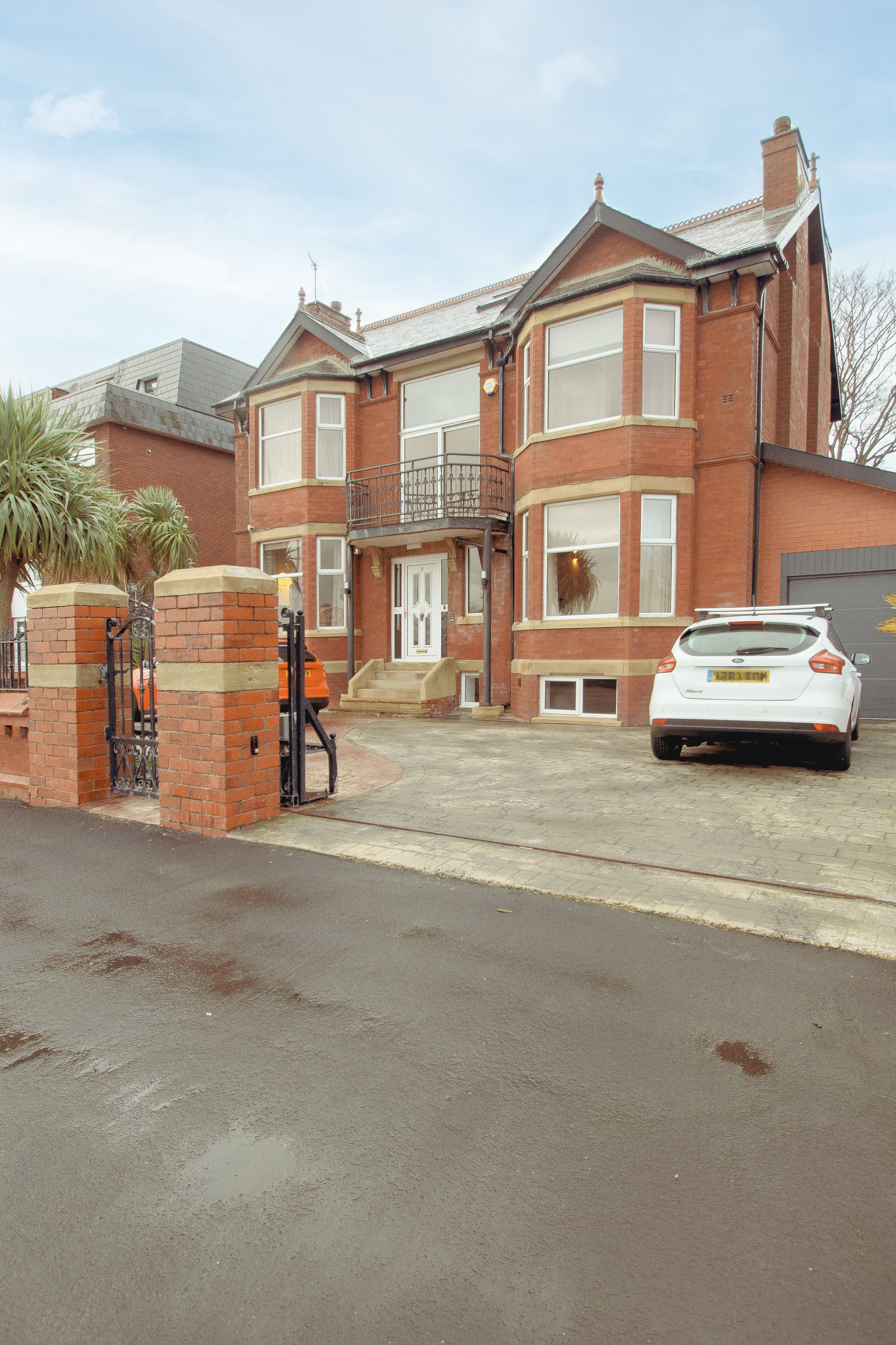 A brick house with large windows, a small balcony, aand a driveway with cars.