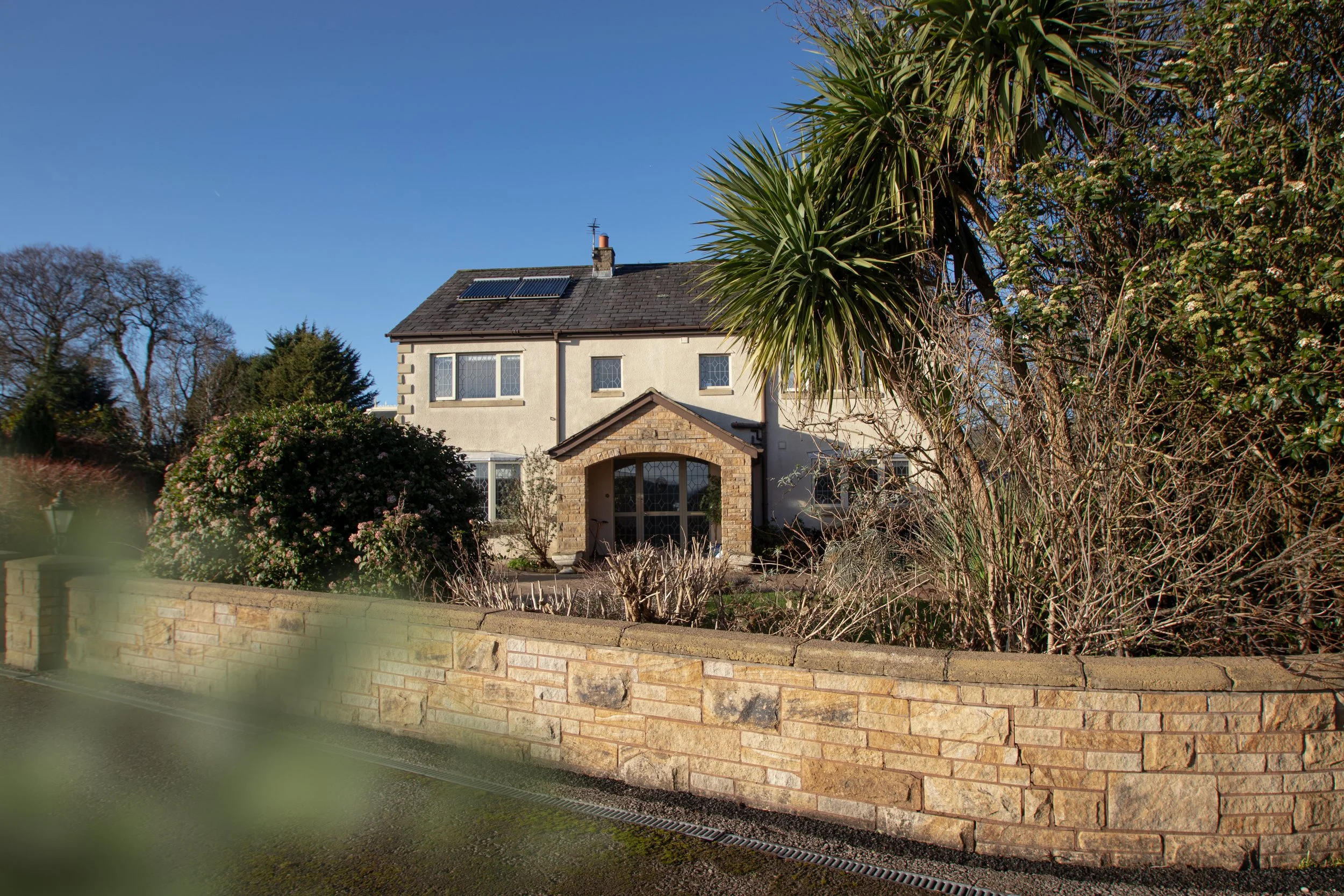 A house with a gray roof, solar panels, and a stone archway over the front door, surrounded by trees, bushes, and a yellow brick wall in the foreground, under a clear blue sky.