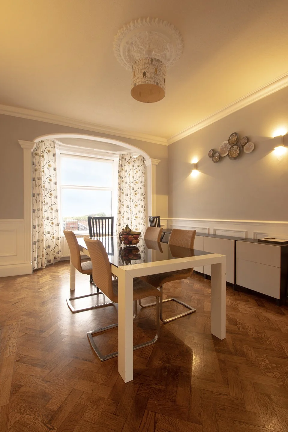 Dining room with a glass table, six chairs, window with floral curtains, wood flooring, wall decorations, and ceiling light fixture.