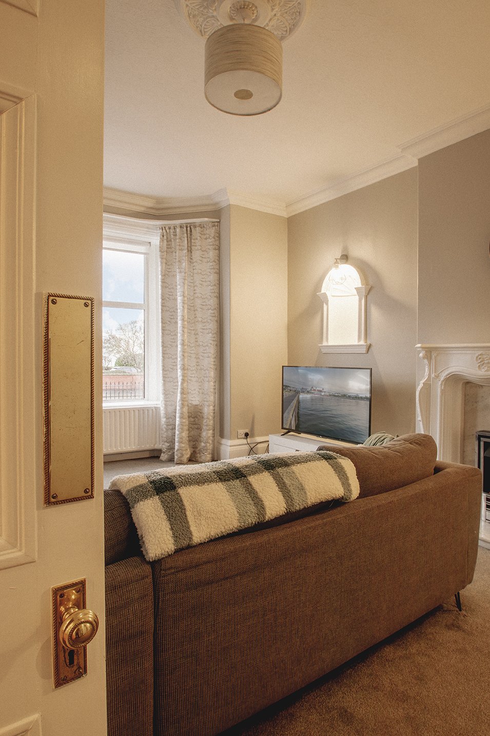 Living room with beige walls, a brown sofa with a plaid throw blanket, a white media console with a TV, decorative wall sconces, and a window with patterned curtains.