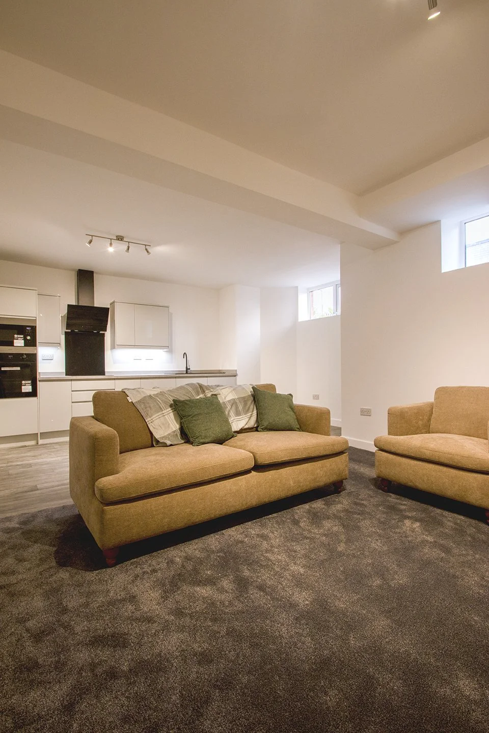 Living room with beige sofas, green pillows, and a view of the kitchen with white cabinets and black accents.