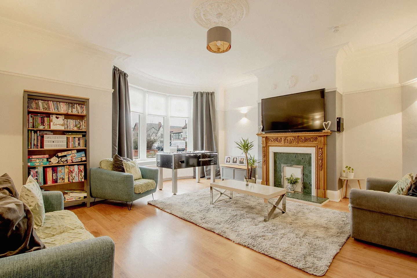 Bright living room with beige walls, large bay window, gray curtains, and hardwood flooring. Contains sofas, a coffee table, a bookshelf, a TV above a decorative fireplace with a green marble surround, and a foosball table.