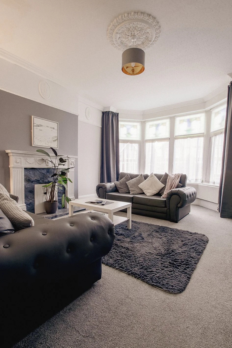 A living room with two gray sofas, a white coffee table, a fireplace with a plant, and large bay windows with curtains, decorated in neutral tones.
