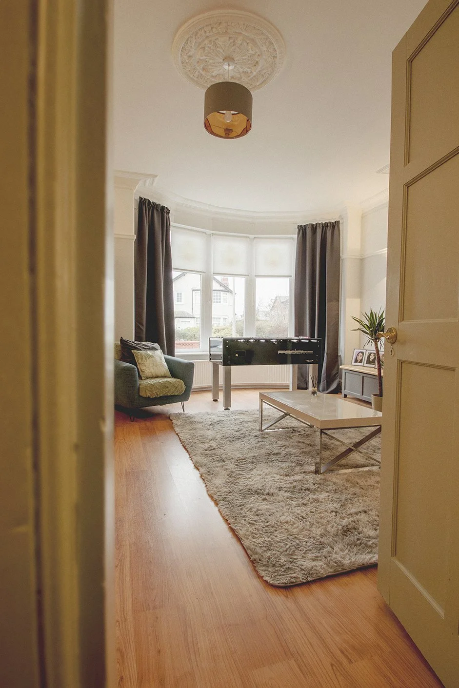 Living room with bay window, dark curtains, gray sofa with cushions, white shaggy rug, white coffee table, TV on a stand, potted plant, and framed photos, viewed from doorway.