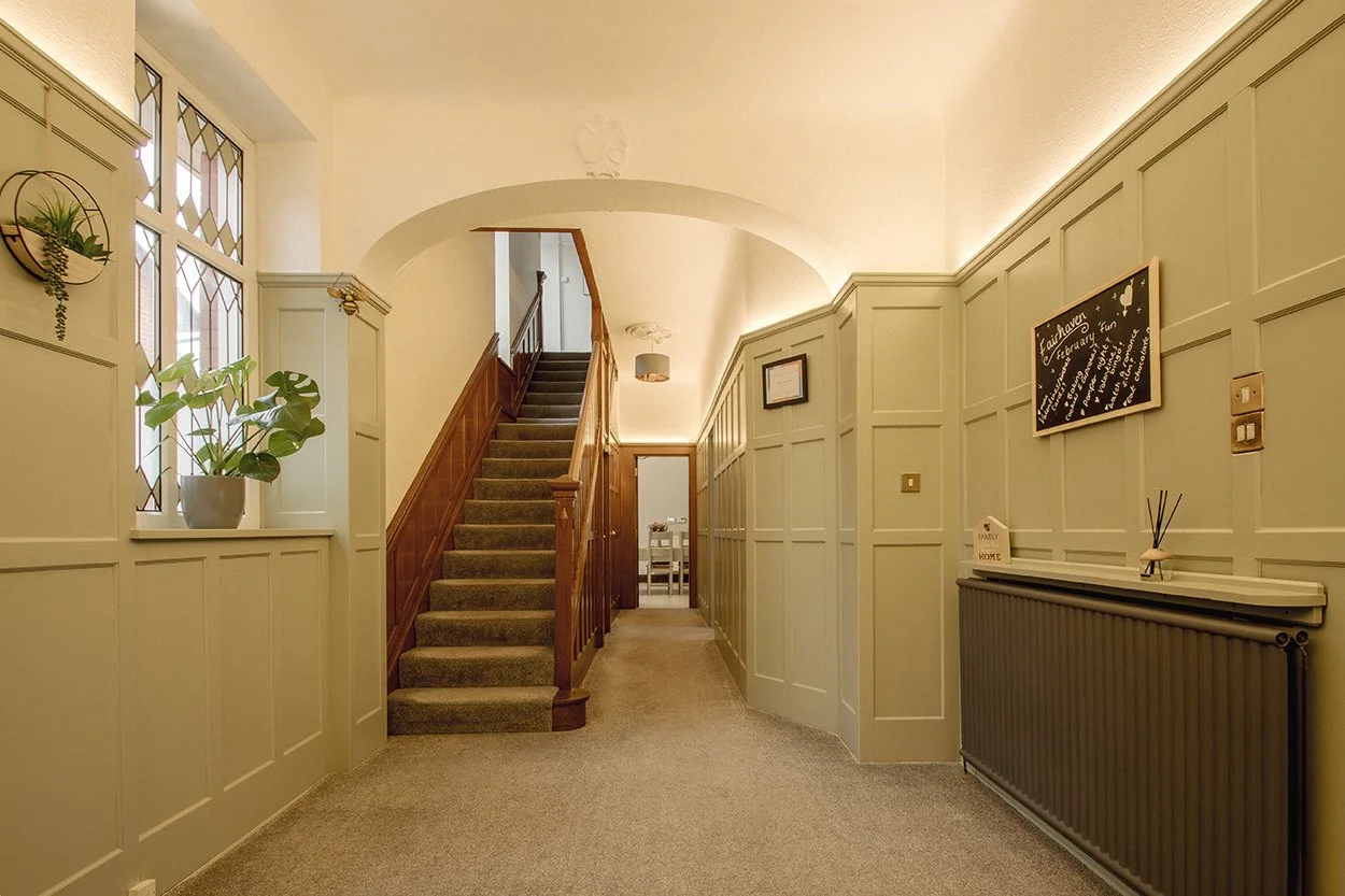 A hallway with beige walls, carpeted floor, a staircase with a wooden handrail, a window with stained glass, a potted plant, a radiator, and a blackboard with handwritten notes.
