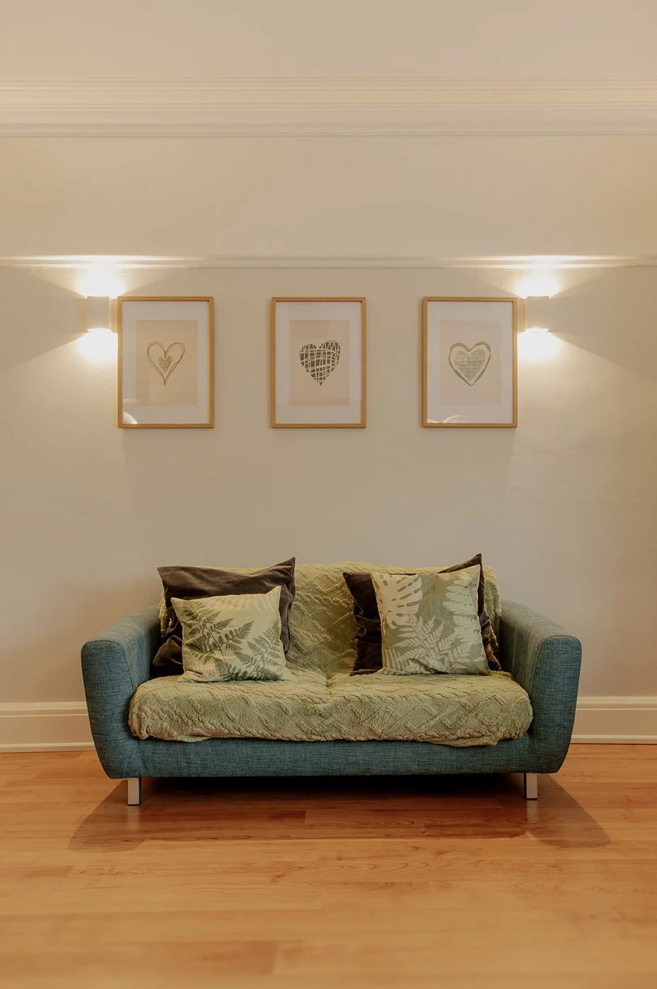 A teal sofa with four pillows, two with leaf patterns and two in dark solid colors, in front of a white wall decorated with three framed botanical heart illustrations and two wall sconces with warm light.