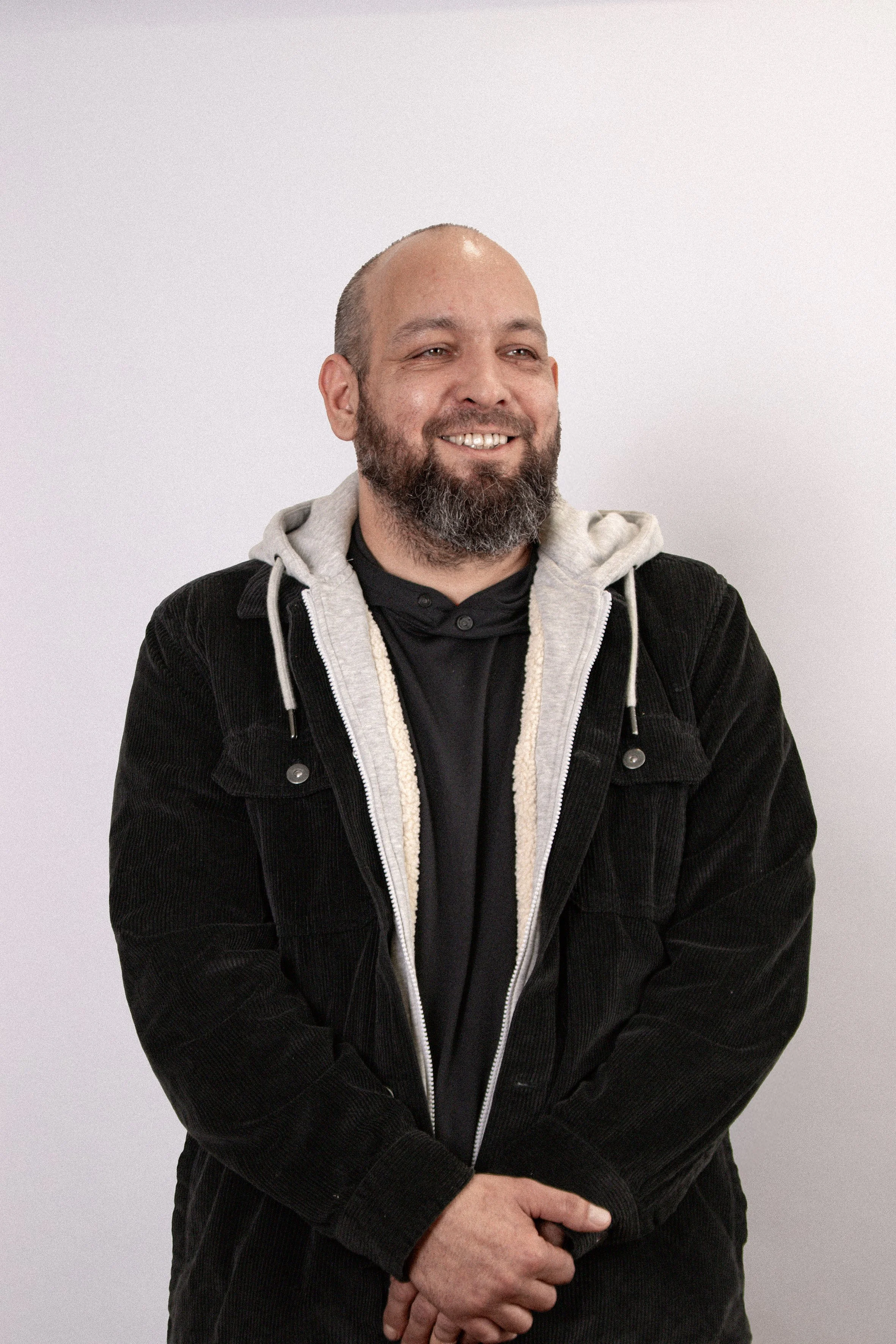 A bearded man wearing a black shirt, gray hoodie, and black jacket standing against a plain white background.