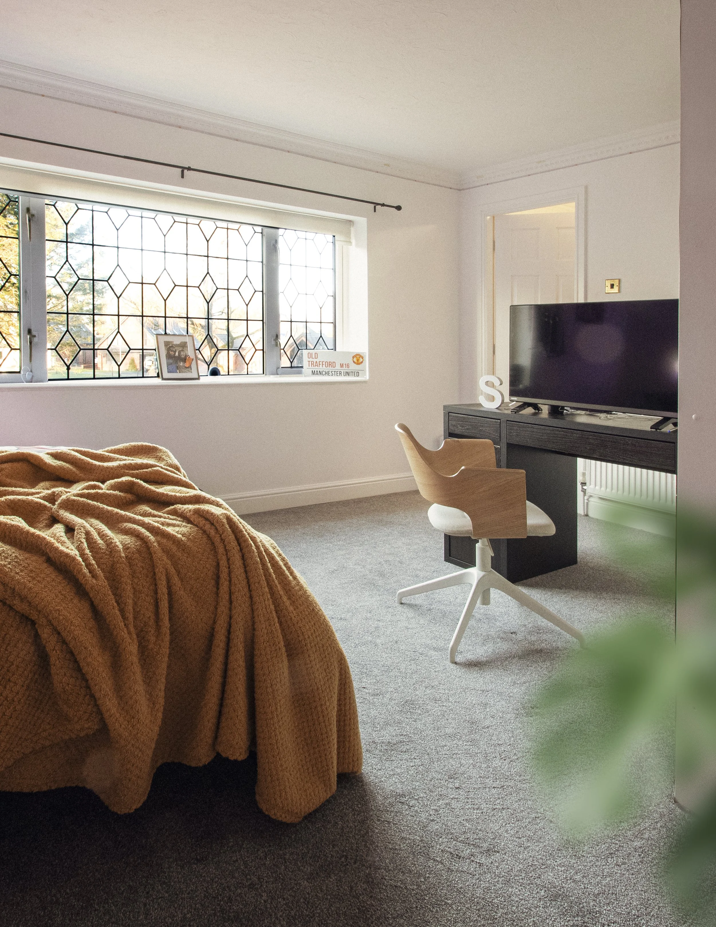 A cozy living room with a beige sofa covered with a brown blanket, a window with decorative black metal grille, a small black speaker, a framed photo, a Manchester United sign, a computer desk with a large flat-screen TV, a wooden desk chair, and a light-colored wall with a doorway.