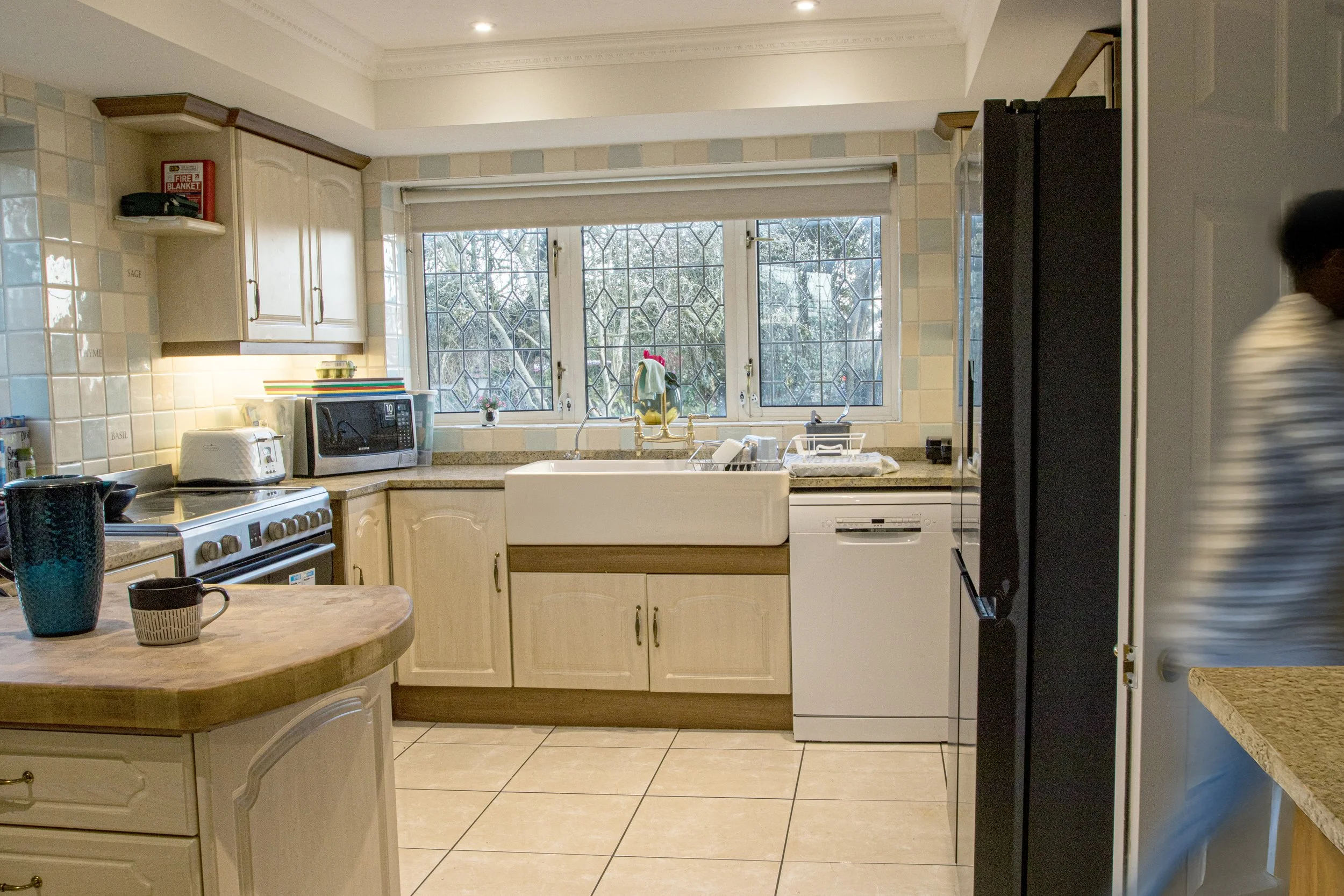 A kitchen with white cabinets, a large window above a farmhouse sink, a countertop with small appliances, and a tiled backsplash.