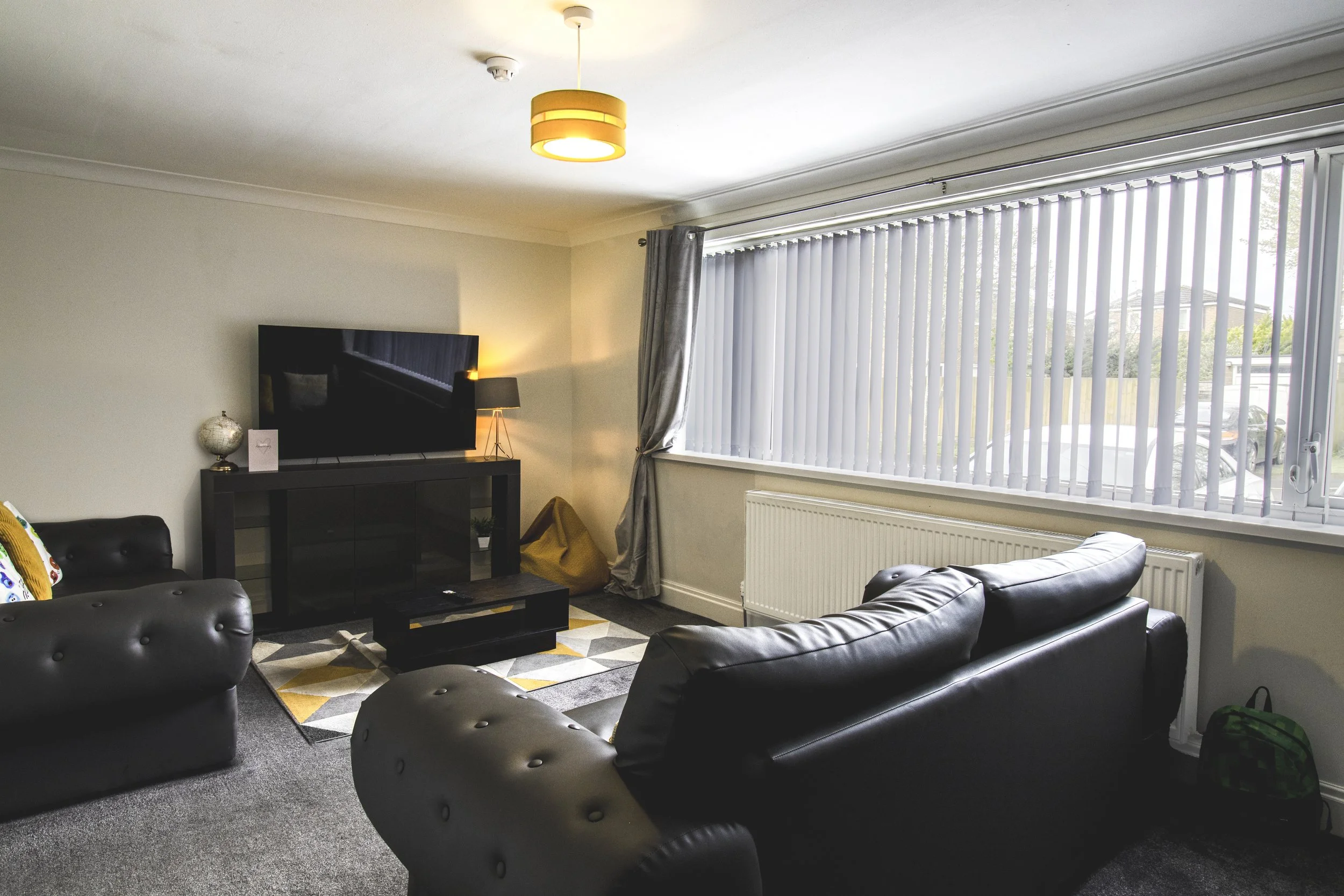 Living room with black leather couches, a flat-screen TV on a black entertainment unit, a window with vertical blinds, a gray curtain, a gray carpet, and a yellow and white geometric rug.