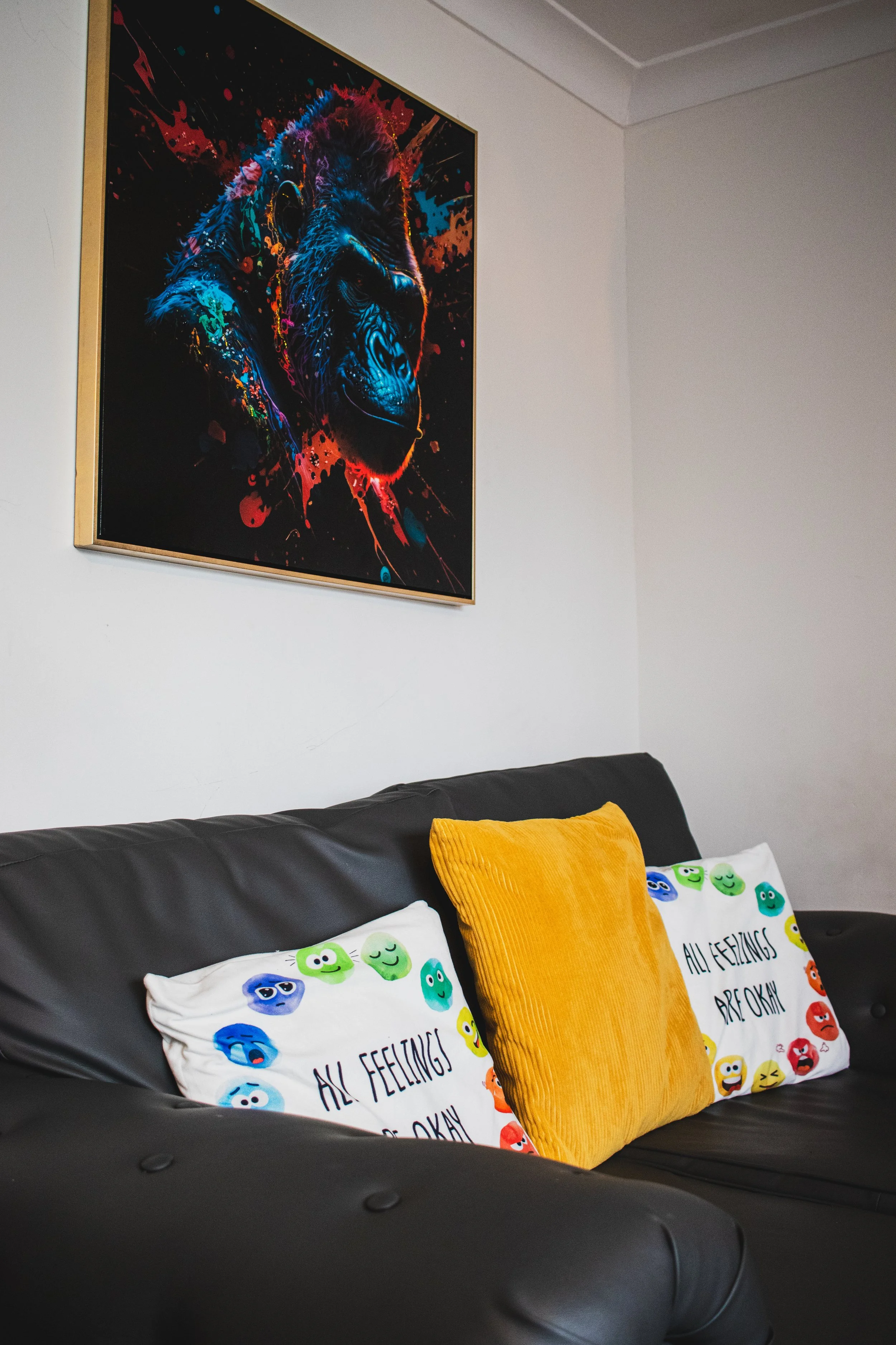 A black leather sofa with three decorative pillows, two featuring colorful emoji faces and one in solid yellow, in a room with white walls. A vibrant, abstract gorilla painting with splashes of bright colors hangs on the wall above the sofa.