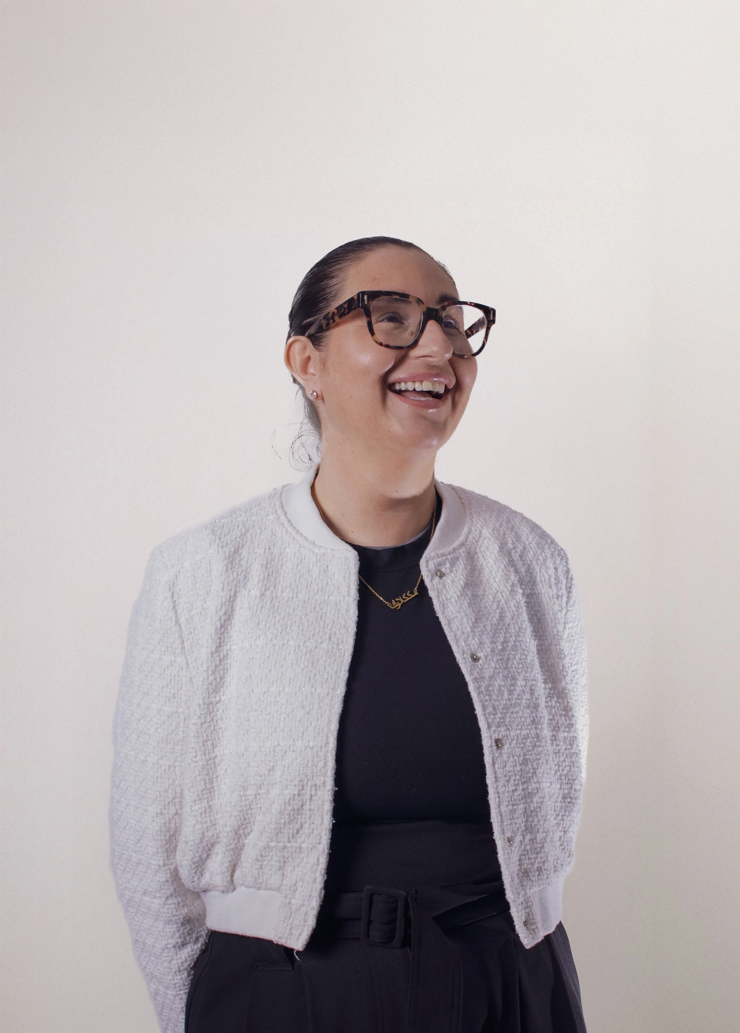 A woman with dark hair pulled back, wearing glasses with tortoise shell frames, a white textured cardigan, a black top, black pants with a belt, and a gold necklace, smiling against a plain white background.