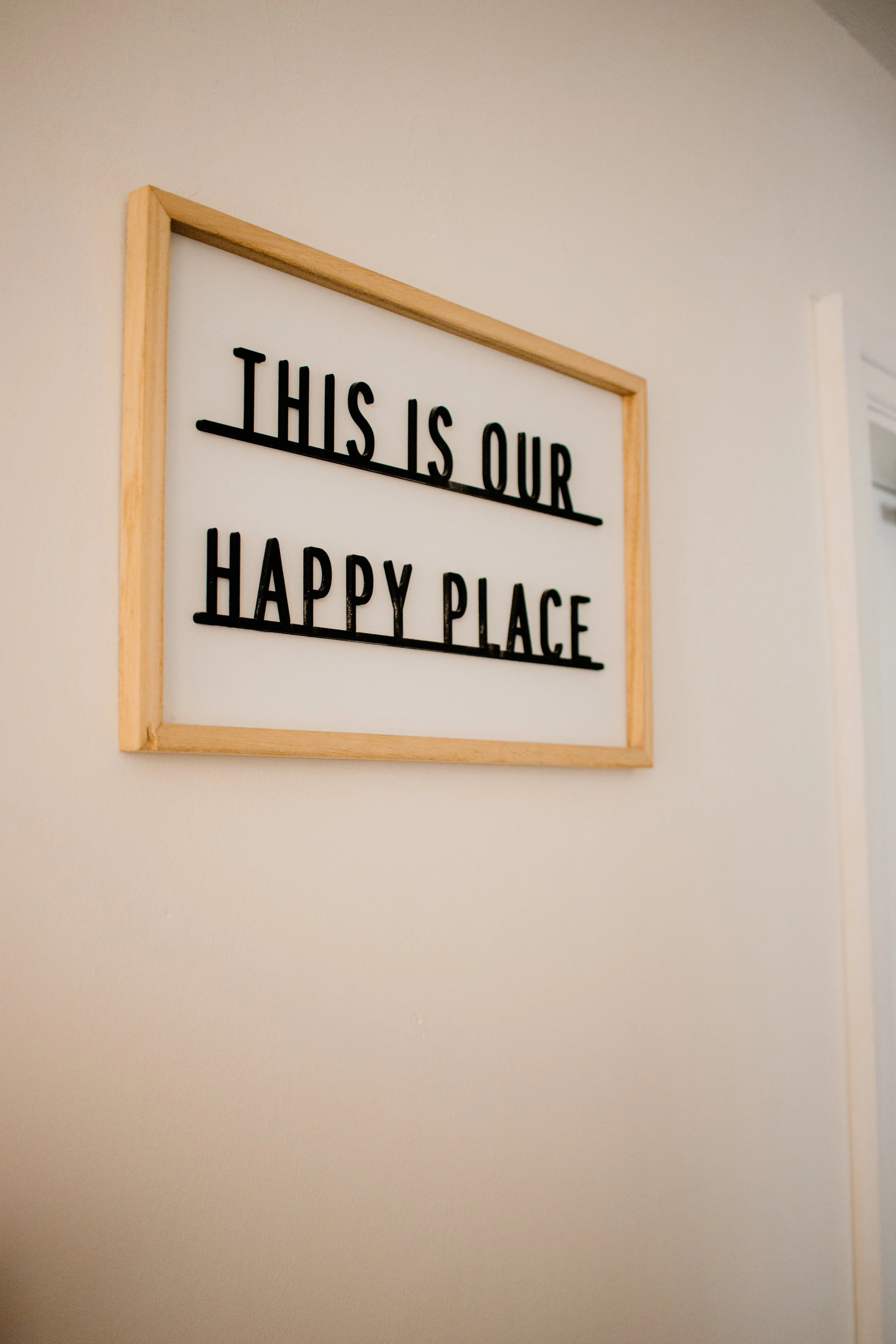 Whiteboard with black text reading 'THIS IS OUR HAPPY PLACE' inside a wooden frame, mounted on a plain wall.