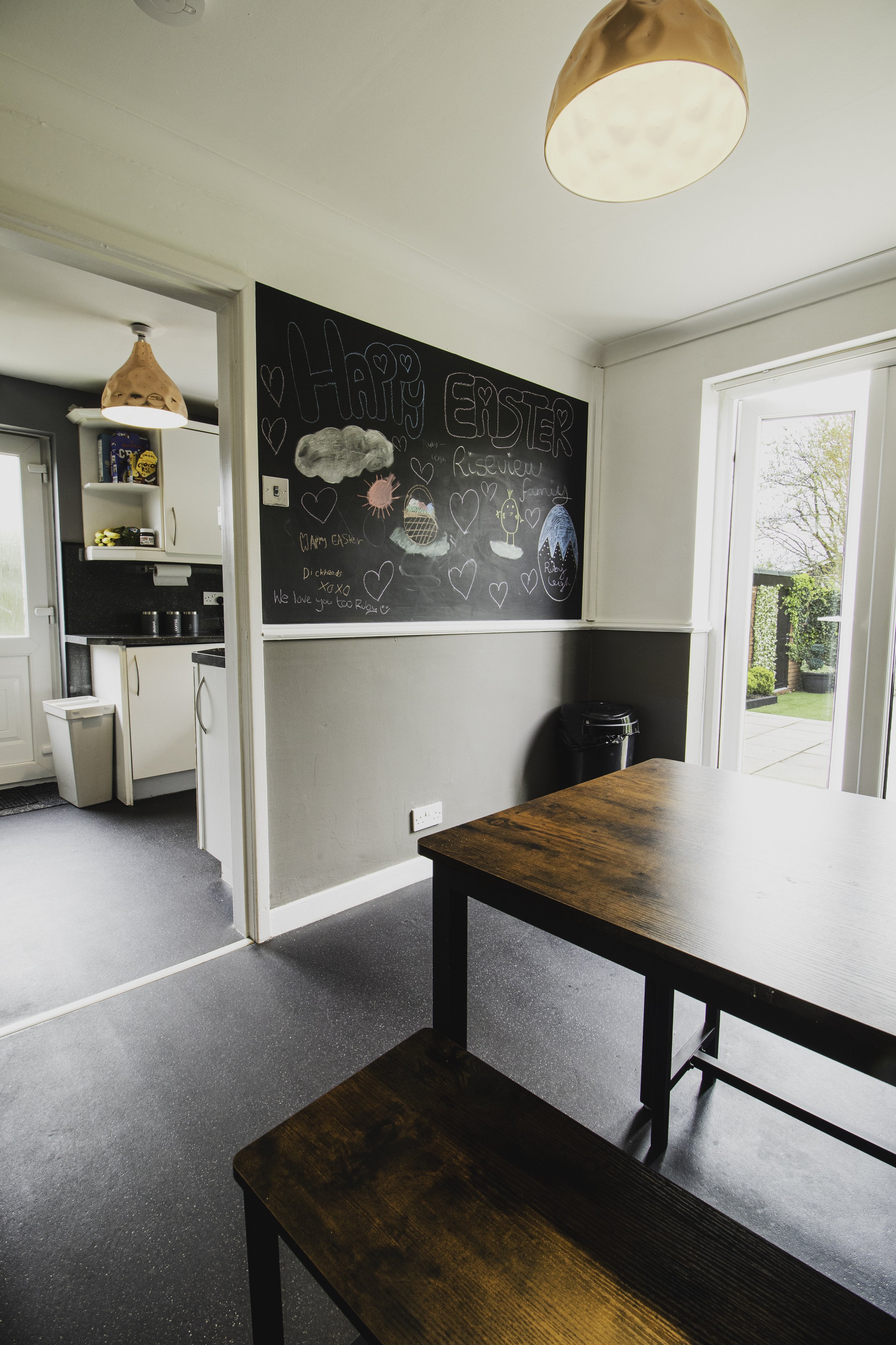 A dining room or kitchen area with a dark wooden table in the foreground, a wall with a chalkboard featuring themed drawings and messages, a window with a view of a garden, and a door leading outside. The ceiling has a copper-colored pendant light.