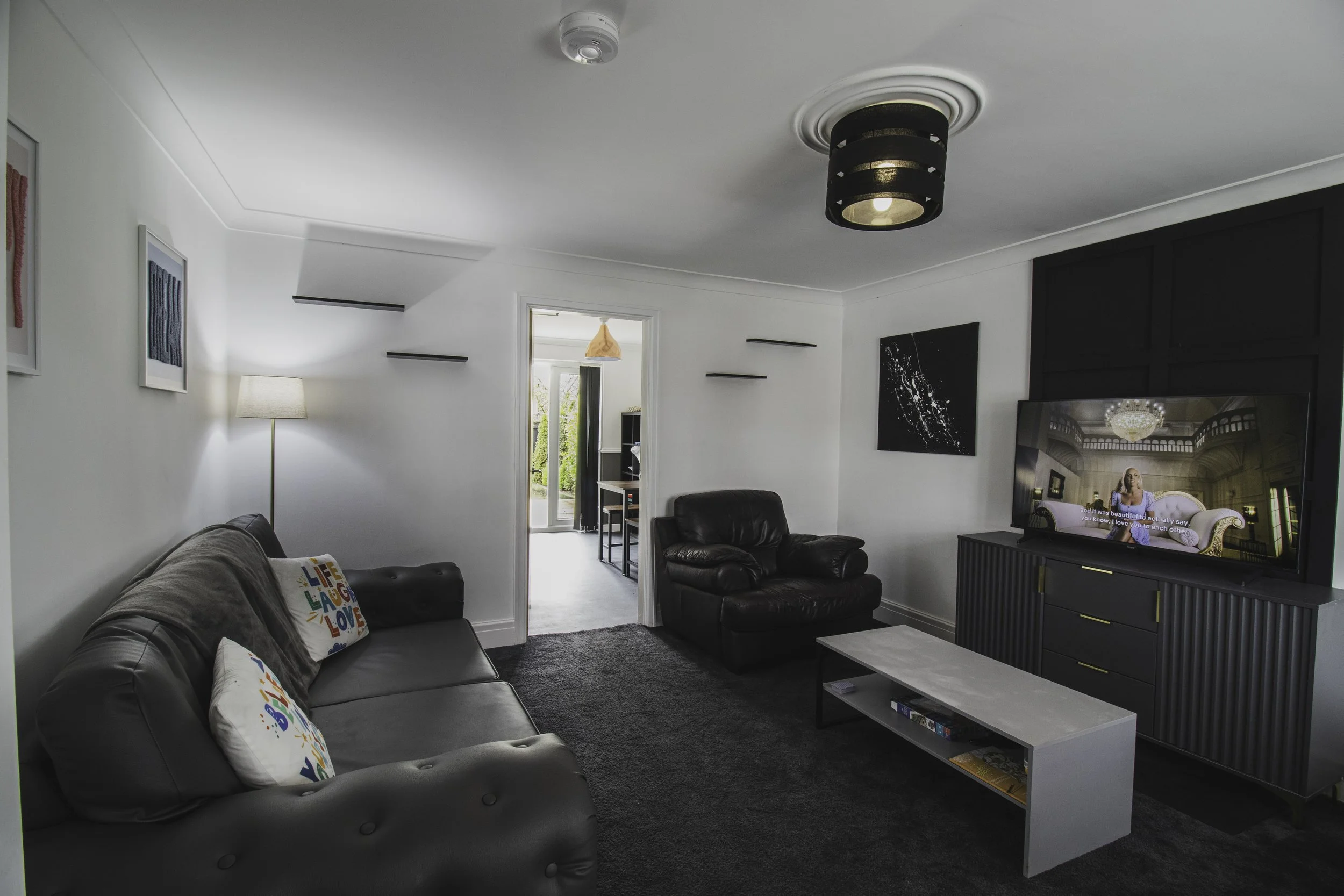 Living room with gray sofa and black leather armchair, white coffee table, large flat-screen TV, and wall art, with a doorway leading outside and a black ceiling light.