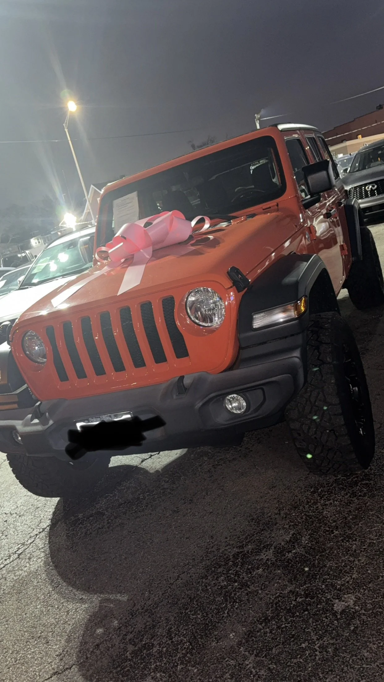 Red Jeep with a pink and white ribbon on the hood, parked in a lot at night.
