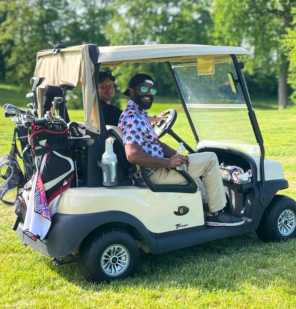 Two men sitting in a golf cart on a golf course, one wearing sunglasses and a Hawaiian shirt, holding a drink. The other man is in the back seat, smiling at the camera. The golf cart has golf clubs attached at the back and a bottle of hand sanitizer hanging from the side. Green trees and grass are in the background.