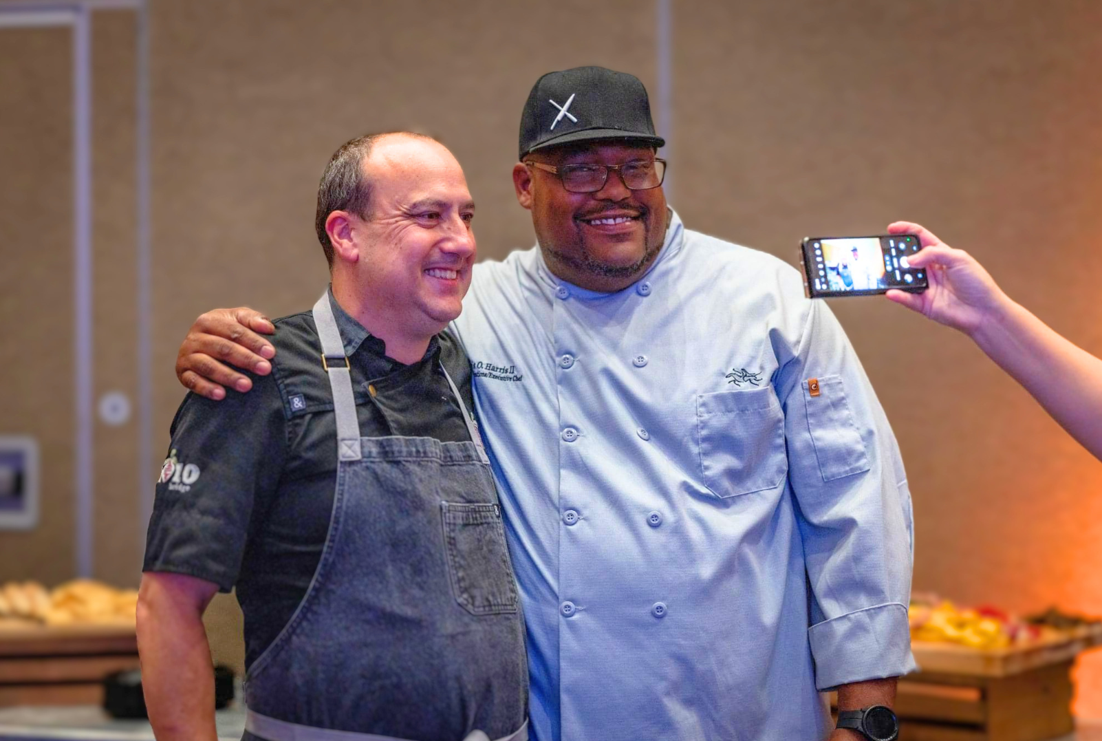Two men pose for a photo, one appears to be a chef in a white coat and the other a man in a black chef uniform. Someone is taking their picture with a smartphone.