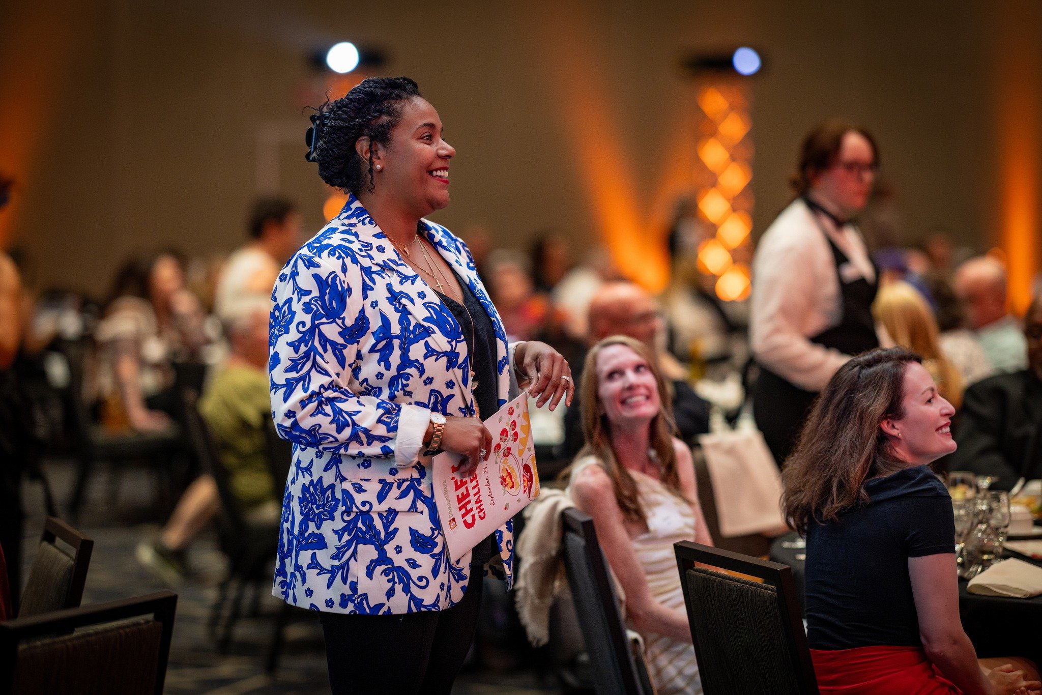 A woman smiling and standing at a seated event, holding a pamphlet, in a large room with an audience.