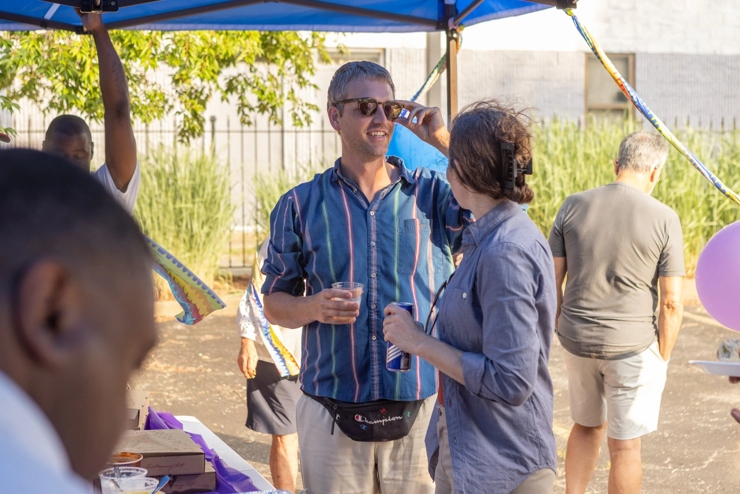 A group of people at an outdoor gathering under a blue canopy, with a man in sunglasses smiling and holding a drink, interacting with a woman with glasses, while others stand nearby and colorful decorations hang from the canopy.