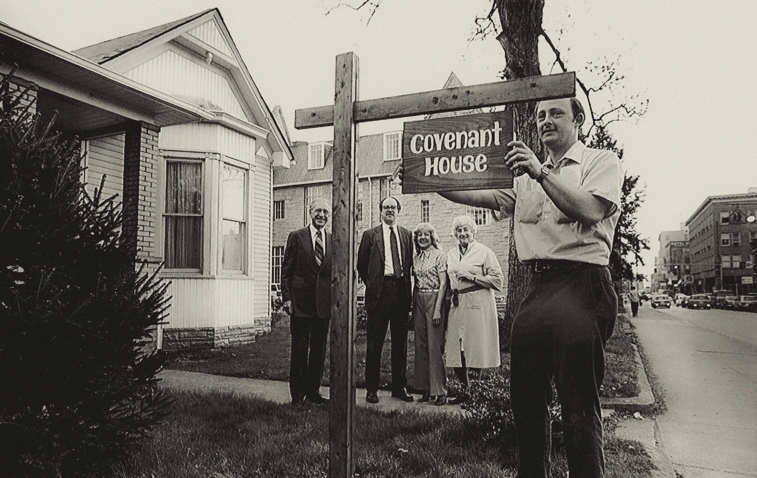 A man holding a sign that reads 'covenant house', standing beside a group of four people, with a building and trees in the background, in black and white.