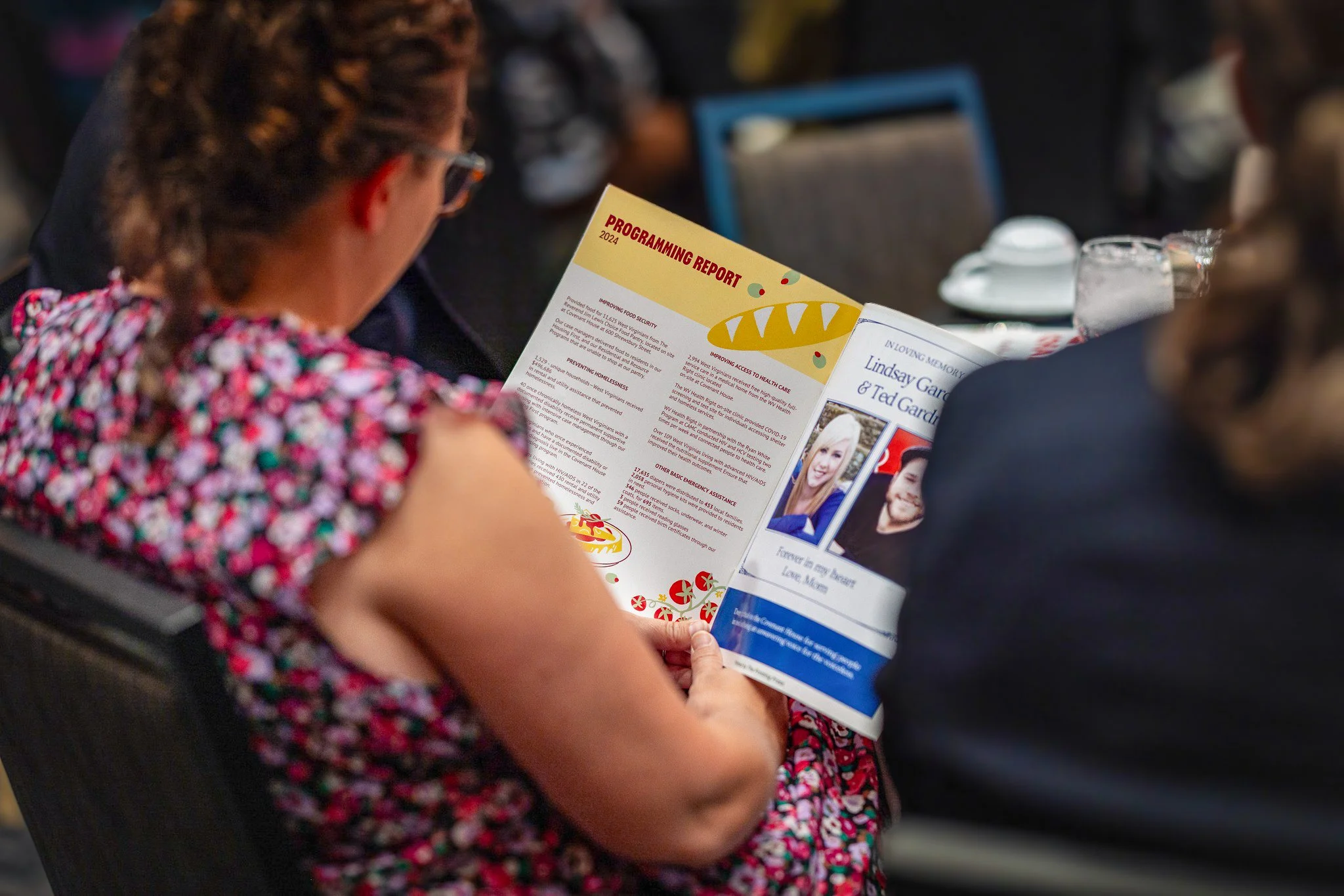 A person with curly hair wearing glasses and a colorful patterned dress reading a program at an event.