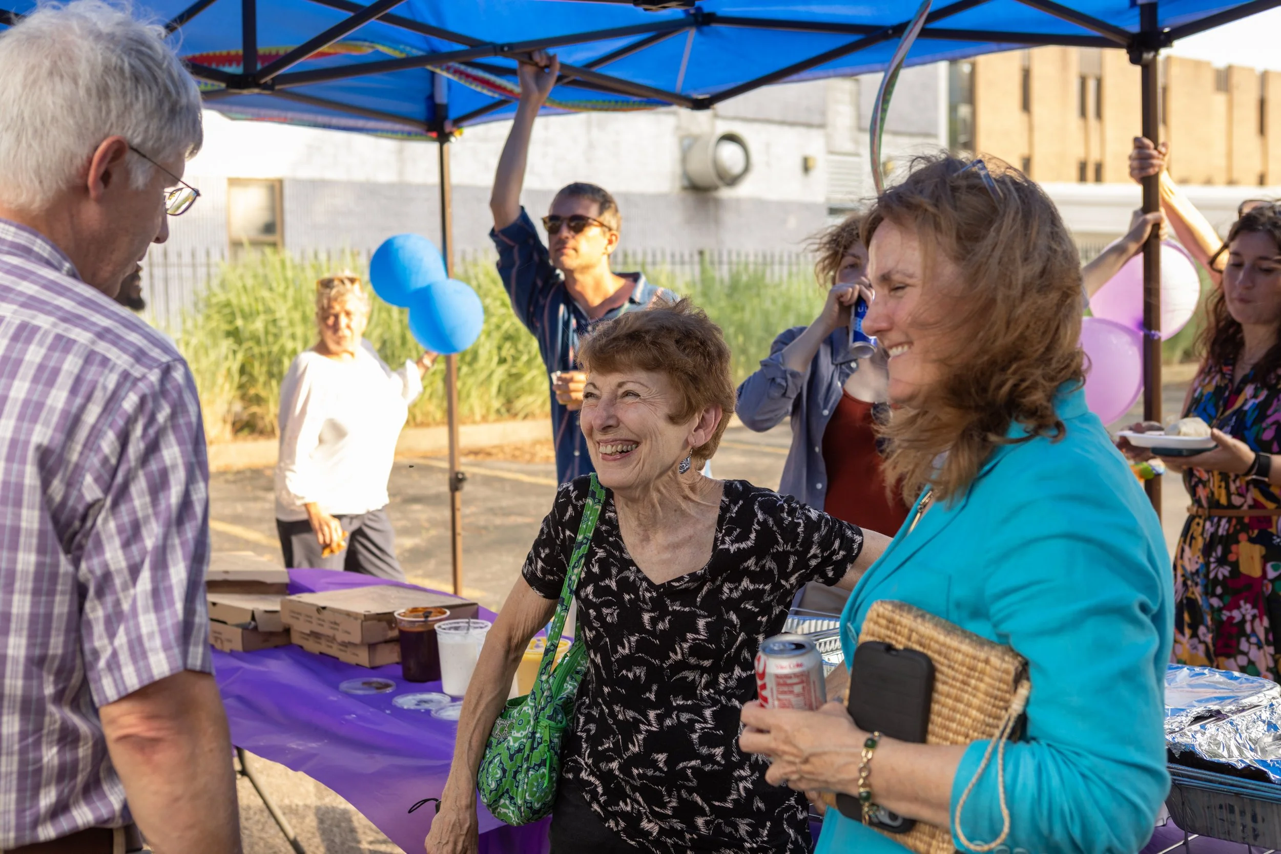 People gathered at an outdoor event, smiling and talking near a table with food and drinks under a blue canopy. A woman in a black and white patterned shirt is smiling and engaging with another woman in a bright blue jacket.