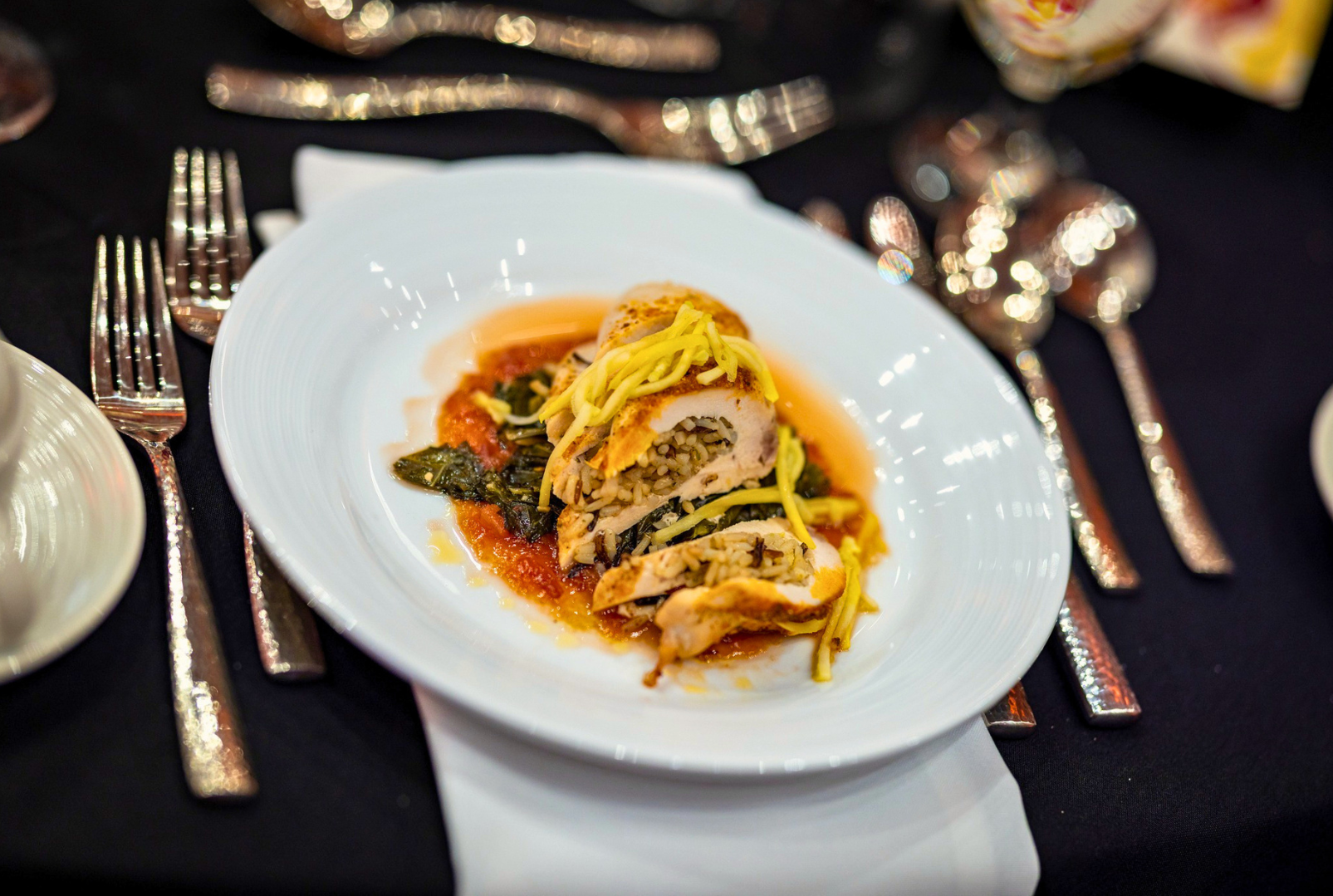 Plate of stuffed chicken breast with rice and spinach, topped with shredded cheese, on a white dish, set on a elegant table with silverware and cutlery.