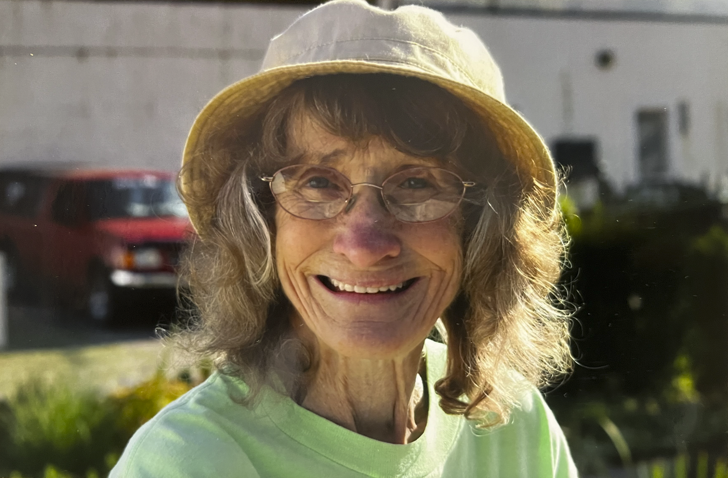Close-up of an elderly woman smiling outdoors, wearing glasses, a yellow hat, and a light green shirt, with a red car and greenery in the background.