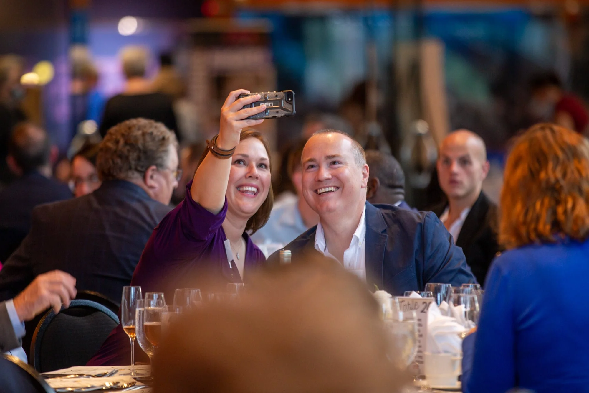 A woman with reddish hair and a man with brown hair sitting at a banquet table, smiling as she takes a selfie with her smartphone. Other people are seated around the table, and the background shows a crowded, well-lit event hall with a blurred crowd.