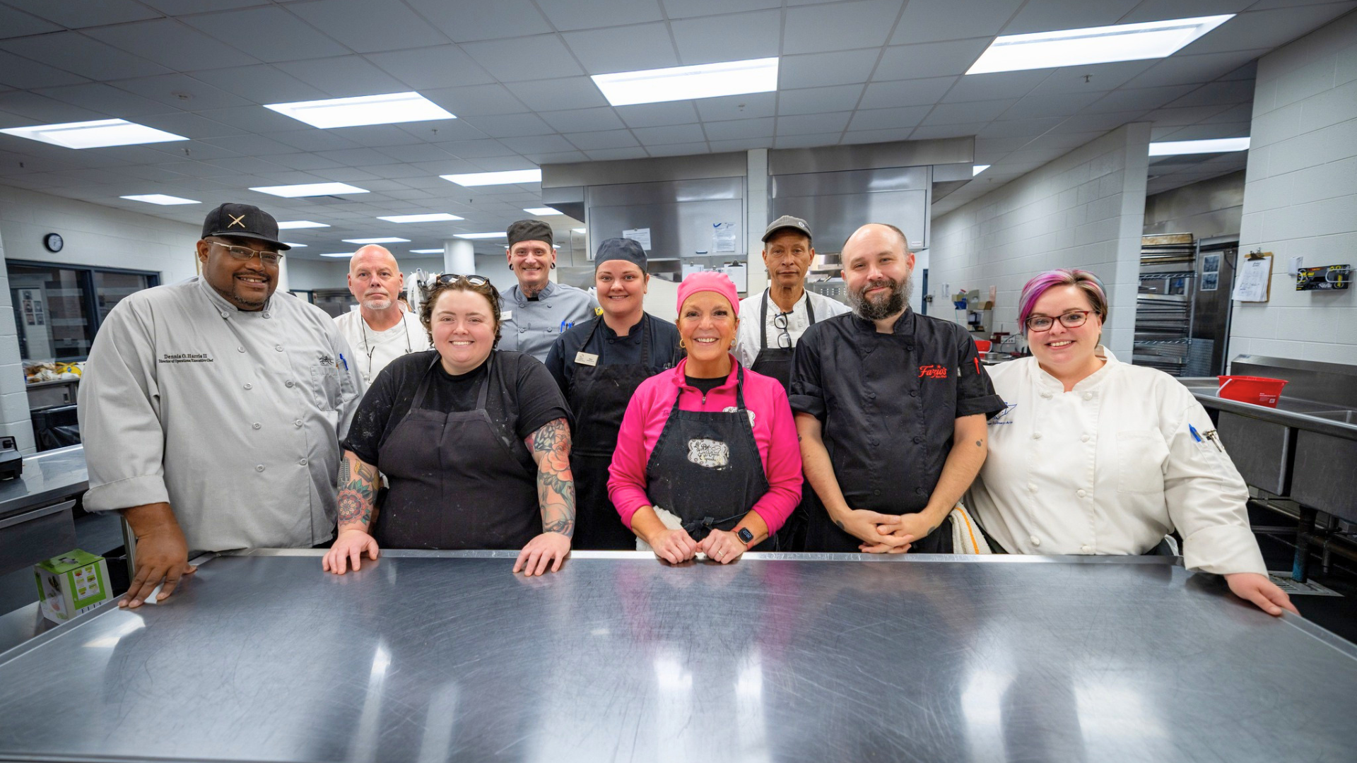 Group of nine diverse chefs and kitchen staff in a commercial kitchen, smiling and posing for the photo.