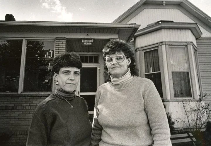Two women standing in front of a house with a sign that says "Covenant House." The women are wearing sweaters and glasses, with short dark hair and slightly longer curly hair, respectively.