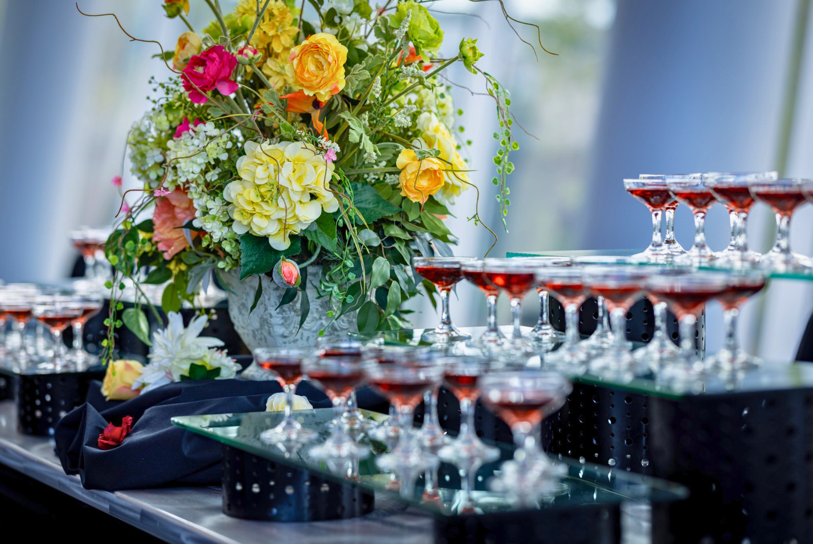 A large floral arrangement with yellow, pink, and white flowers set on a table with multiple small wine glasses filled with red wine, arranged on tiered glass trays.