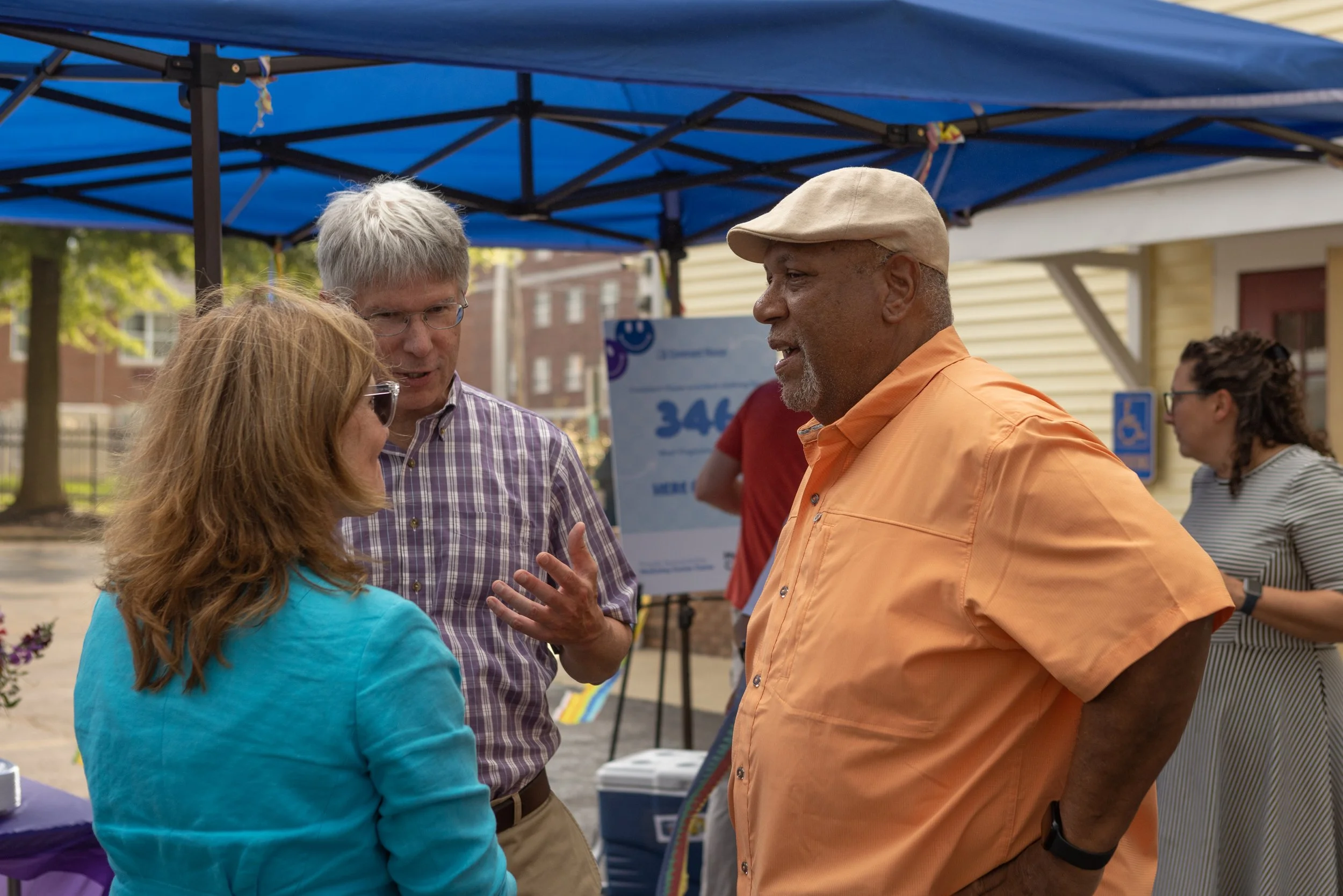 Group of people talking and smiling outdoors under a blue canopy tent, with a yellow house and trees in the background.