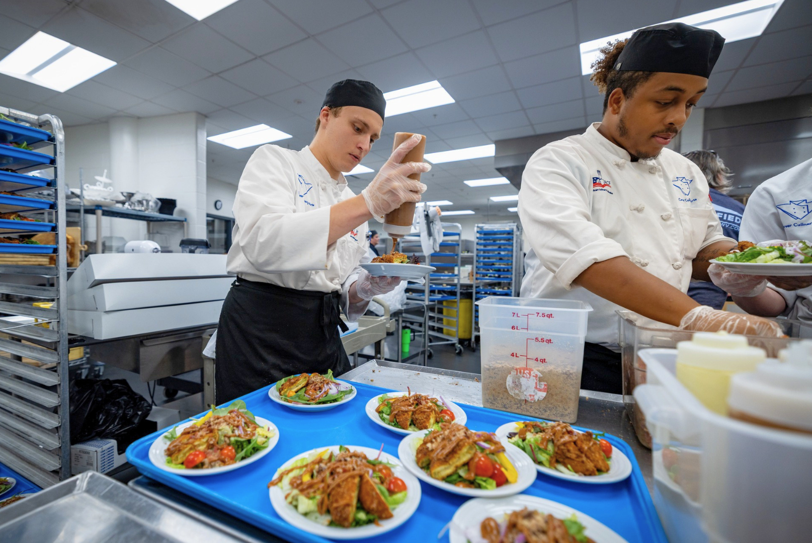 Two chefs in a commercial kitchen preparing plates of salad with fried chicken, cherry tomatoes, and greens, using squeeze bottles for dressing.