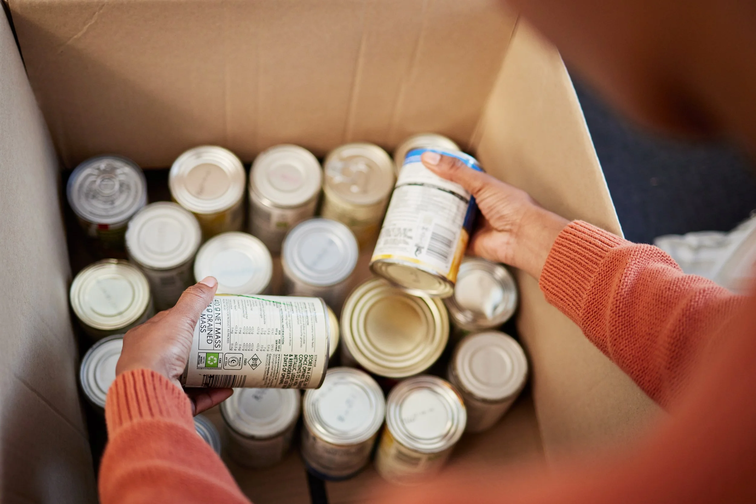 Person in an orange sweater placing canned food into a cardboard box.