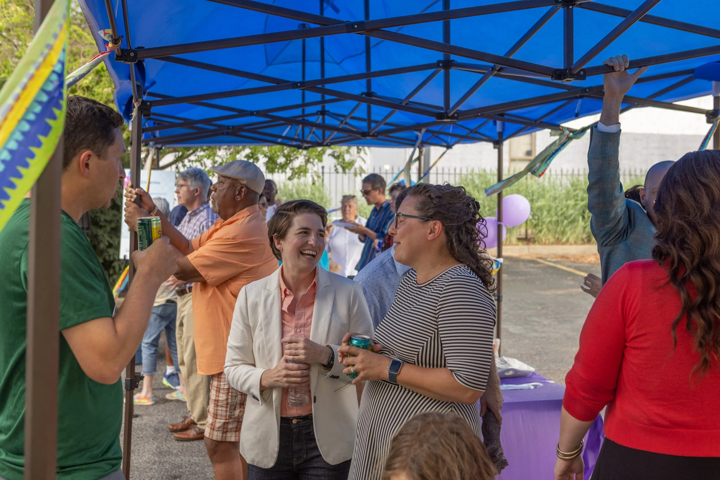 A group of diverse people socializing under a blue canopy at an outdoor event, holding drinks and talking.