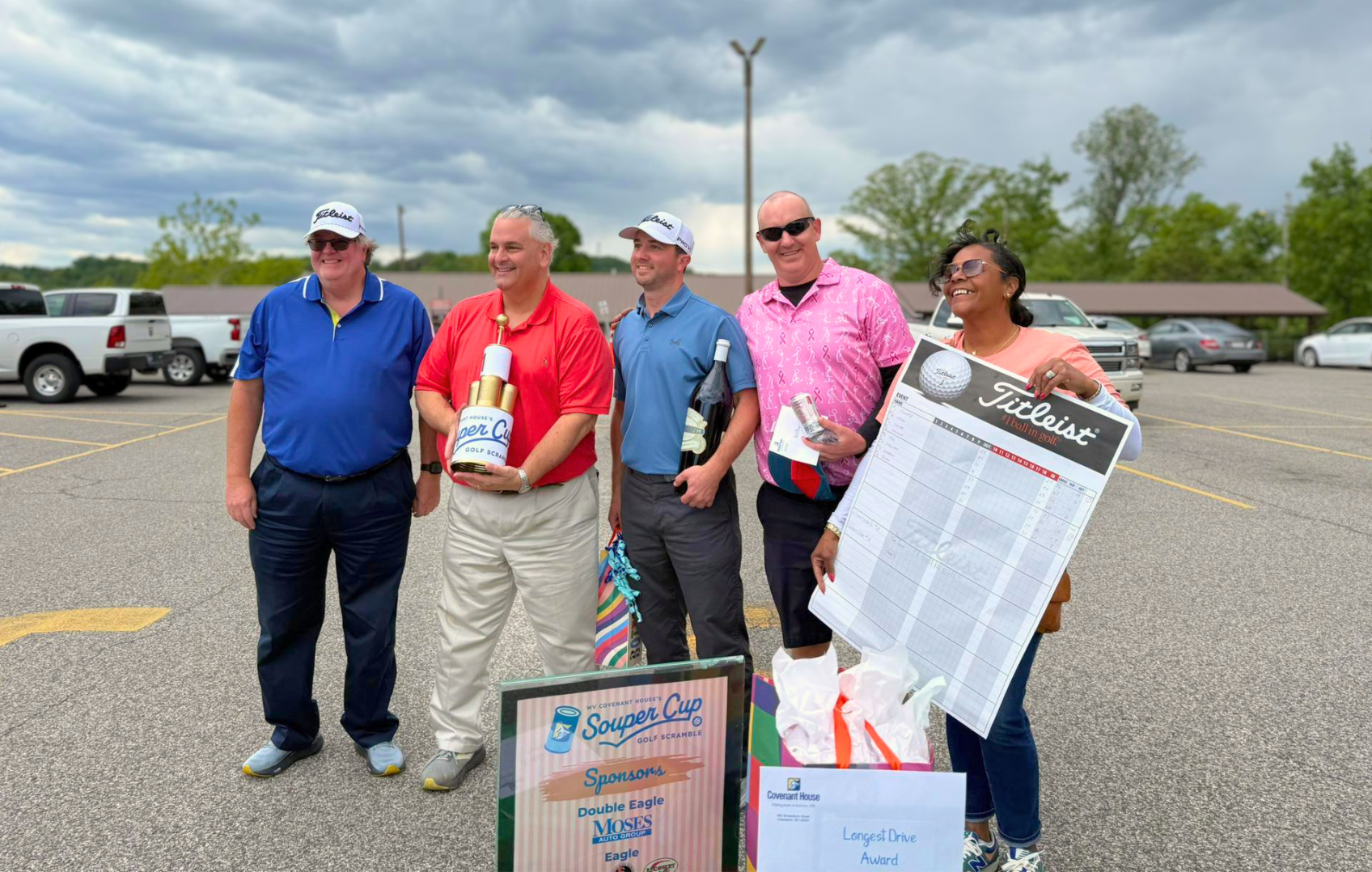Group of five people standing in a parking lot, smiling, during a golf event. One person is holding a large check and a trophy-shaped golf ball. There are several parked cars and trees in the background.