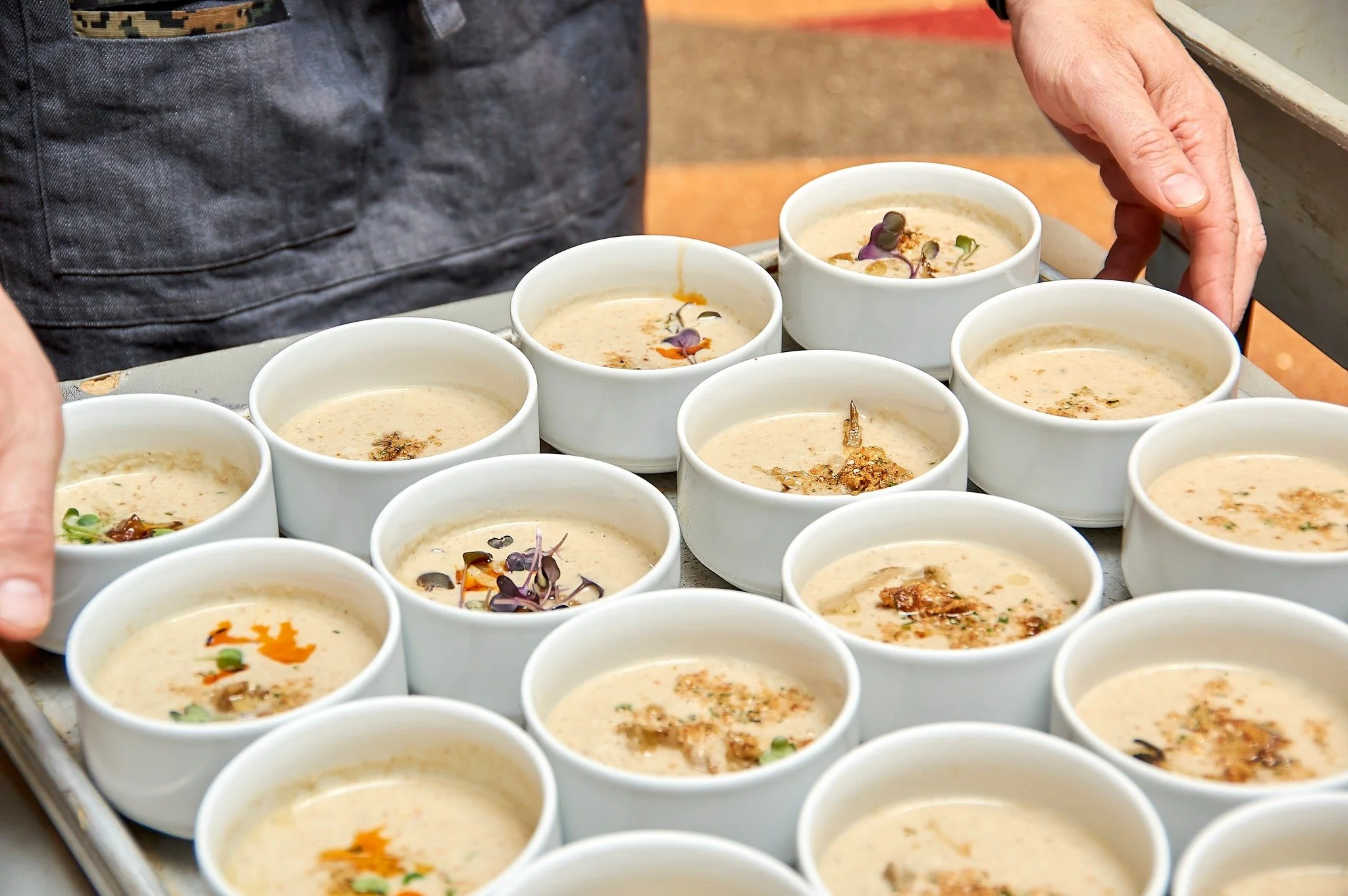 Several small white bowls filled with a creamy soup, garnished with herbs and microgreens, are arranged on a tray. A person with a dark apron is reaching for one of the bowls.