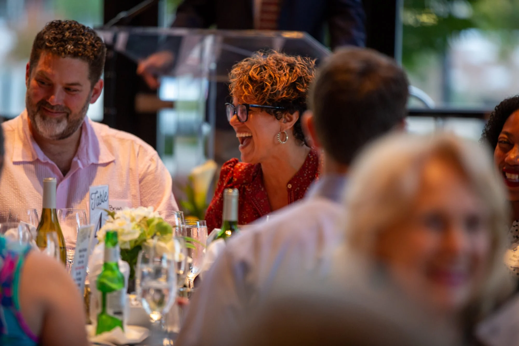 People smiling and engaging in conversation at a dinner table decorated with flowers and wine bottles.