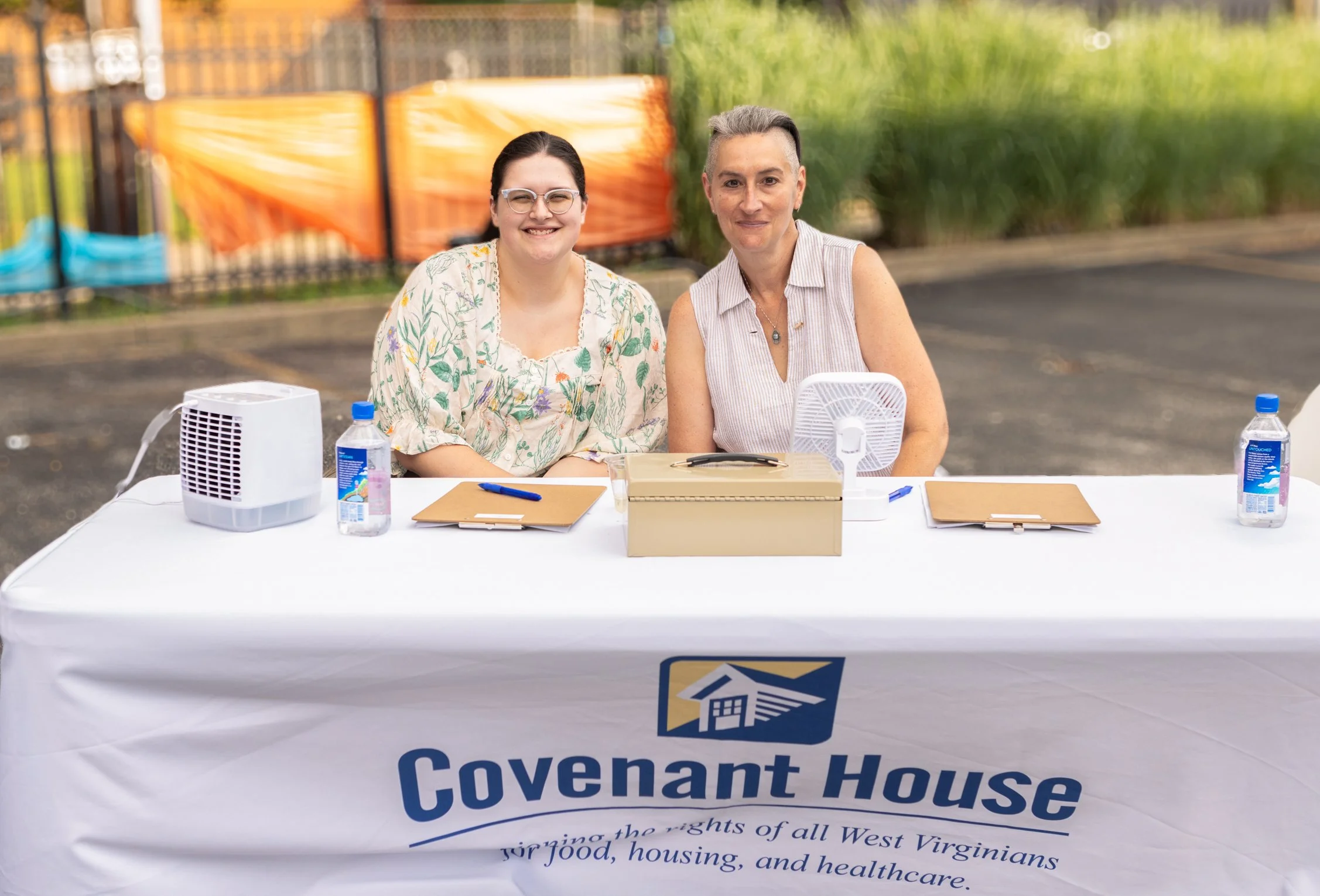 Two women sitting at a table outdoors with a tablecloth displaying the logo of Covenant House. The table has bottled water, a small fan, and clipboards. The women are smiling, and the background includes a fence and green plants.