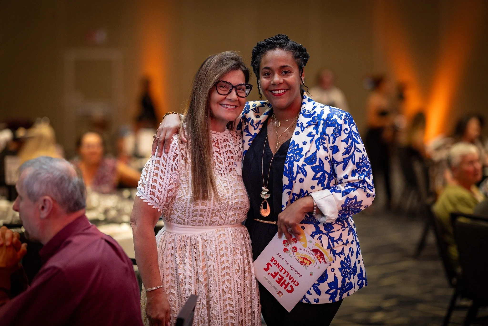 Two women smiling and hugging at an indoor event, one wearing glasses and a white lace dress, the other in a blue and white patterned blazer holding a program. 