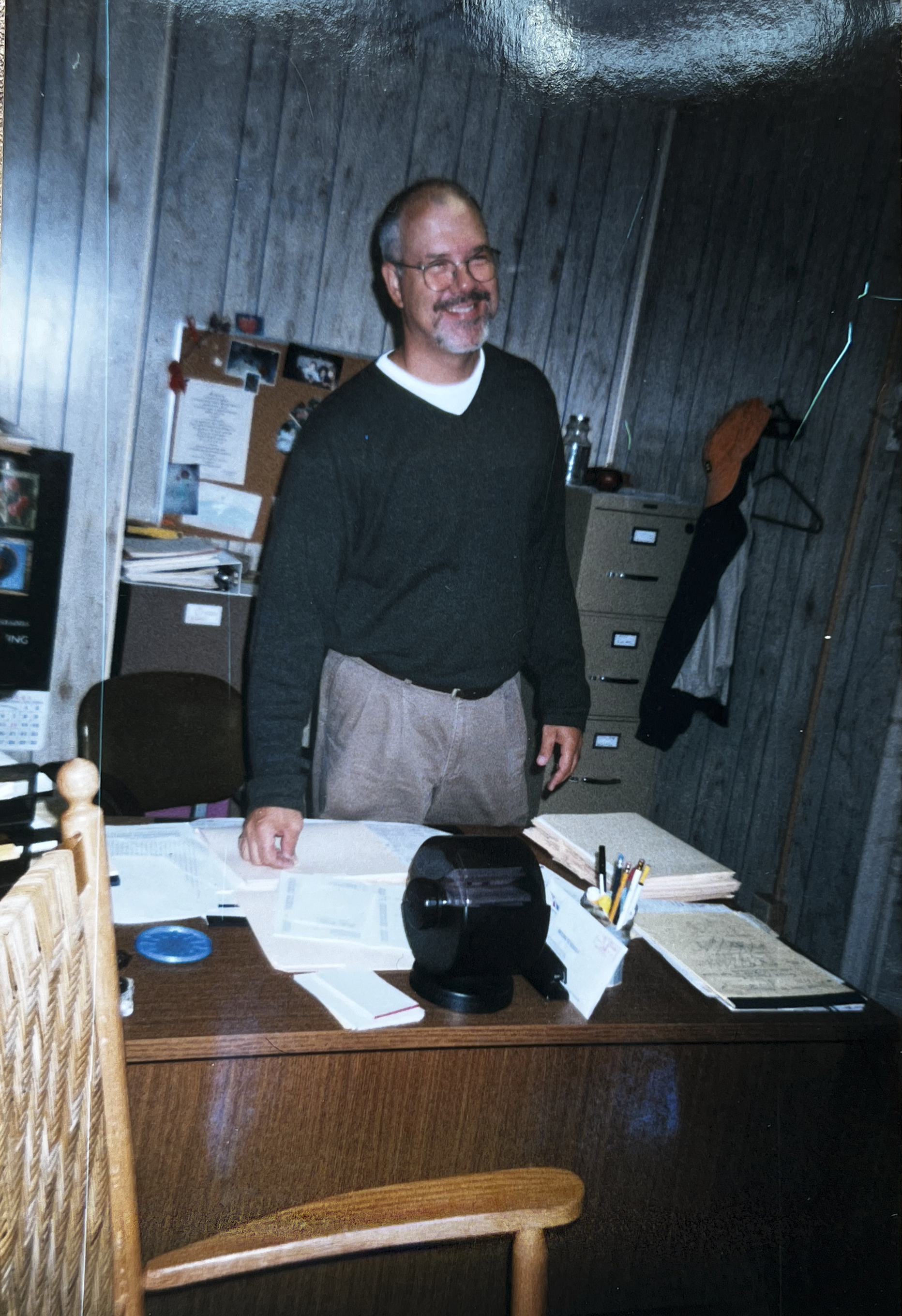 A man with glasses and a beard, wearing a dark sweater over a white shirt, is smiling and standing behind a cluttered wooden desk in an office with wood-paneled walls. There are papers, folders, pens, a telephone, and personal items on the desk. In the background, there's a corkboard with photos and notes, a filing cabinet, a coat hanger with a jacket, and bottles of water.