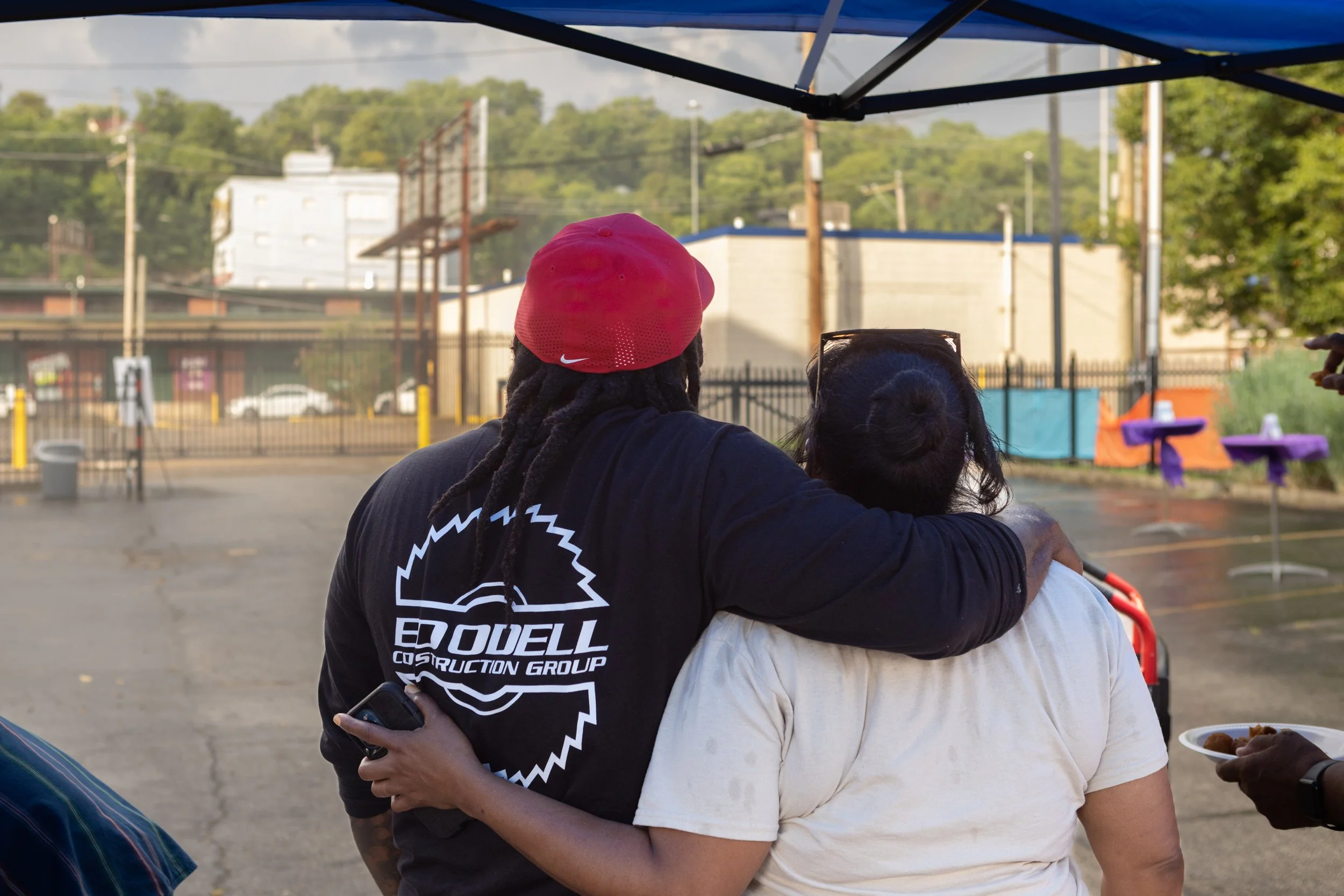 Two people embracing outdoors, one wearing a red cap and a black shirt, and the other wearing a beige shirt, with a blue canopy overhead and a fence, trees, and buildings in the background.