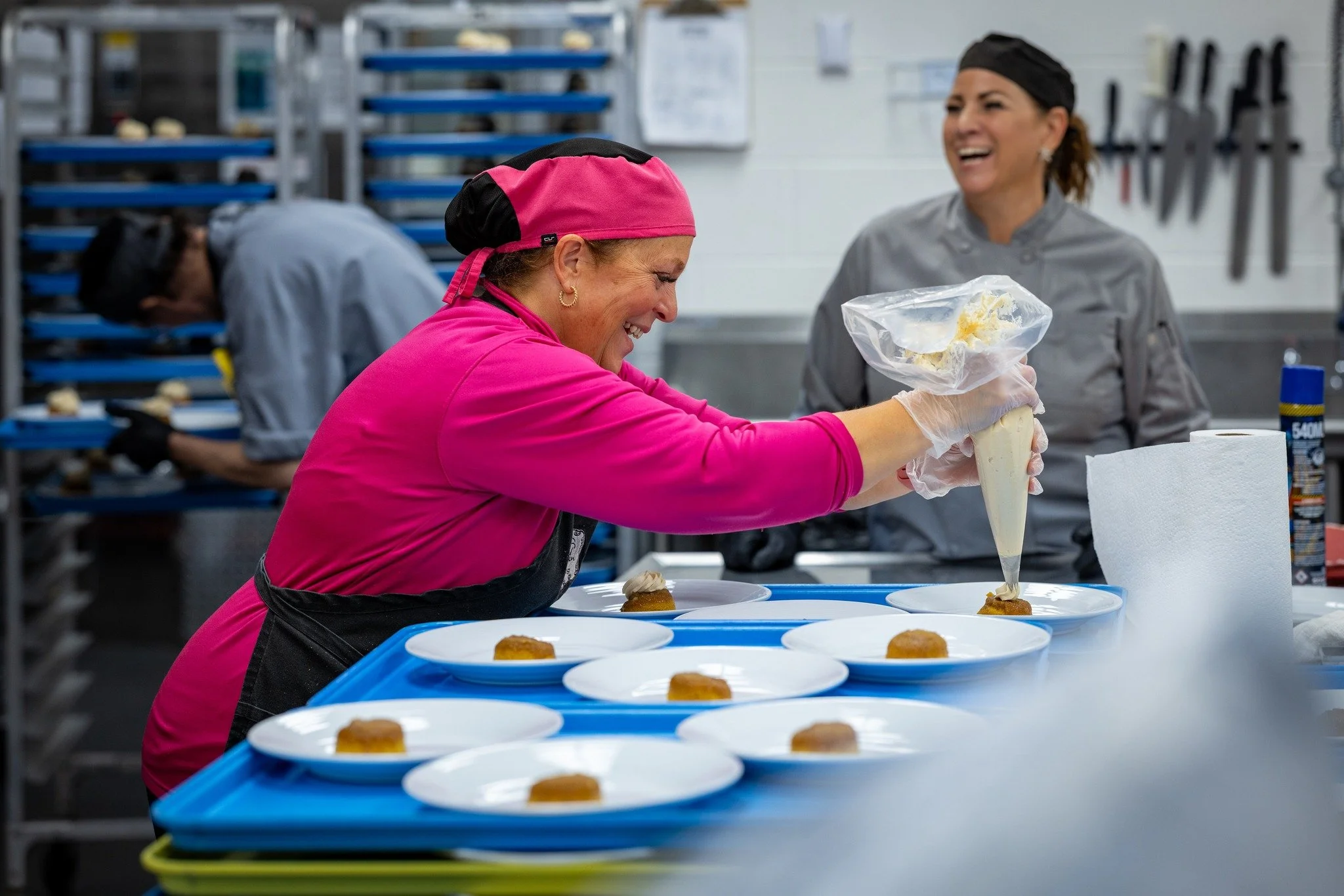 A woman in a pink uniform and black hairnet is piping frosting onto dessert plates while laughing, with another woman in gray uniform and hairnet smiling behind her in a commercial kitchen with racks of baked goods.