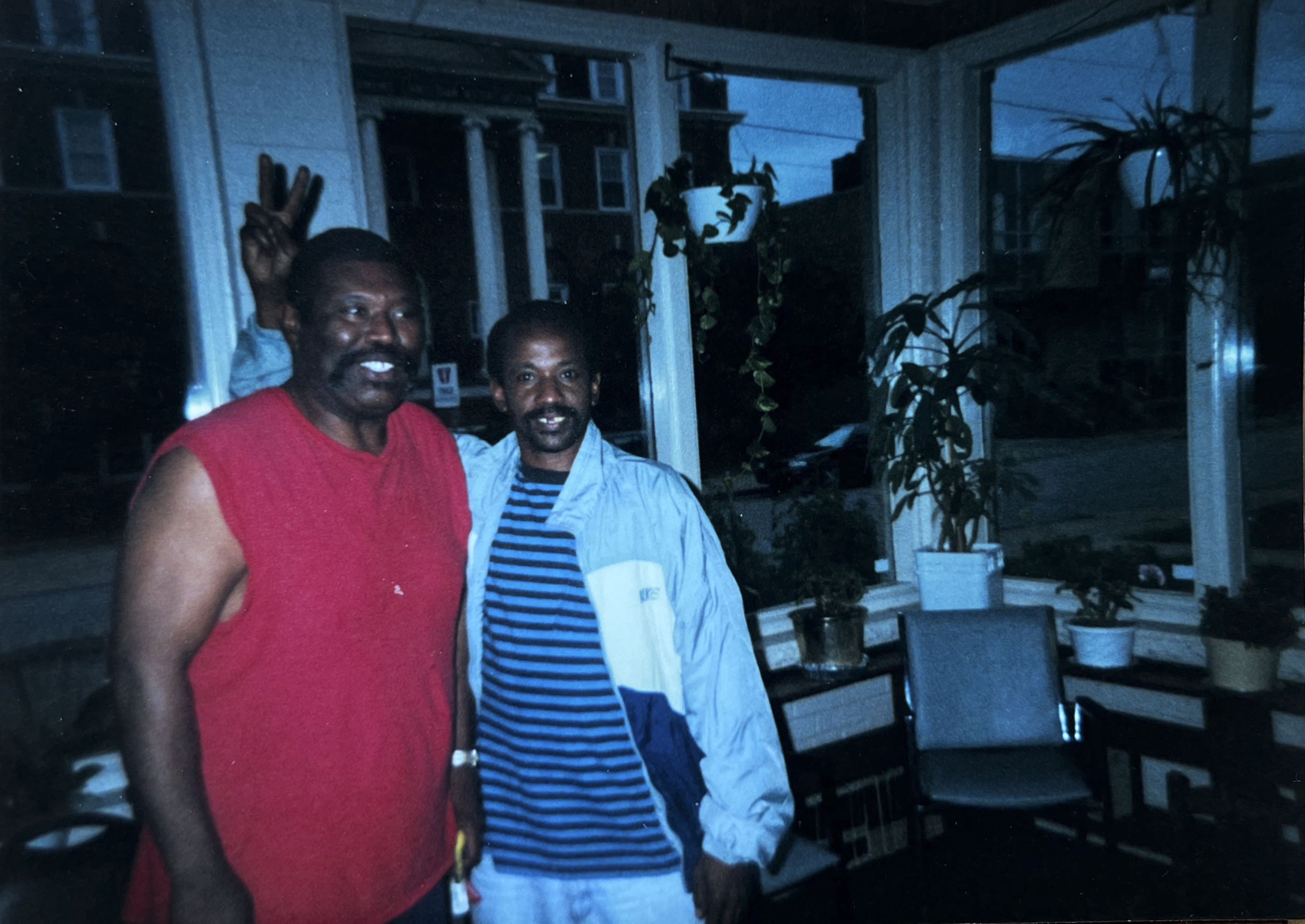 Two men standing inside a house, smiling at the camera. One man is wearing a red sleeveless shirt, and the other is wearing a blue striped shirt and a light jacket. There are large window panes, potted plants, and some furniture in the background, with a dark outdoor scene visible through the windows.